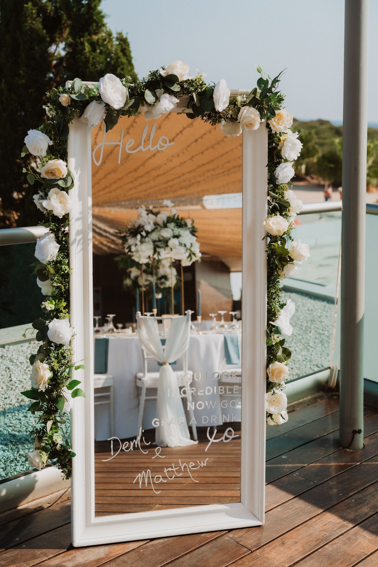 Elegant wedding mirror with white floral garland, greenery, and personalized calligraphy greeting outdoors near a pool.