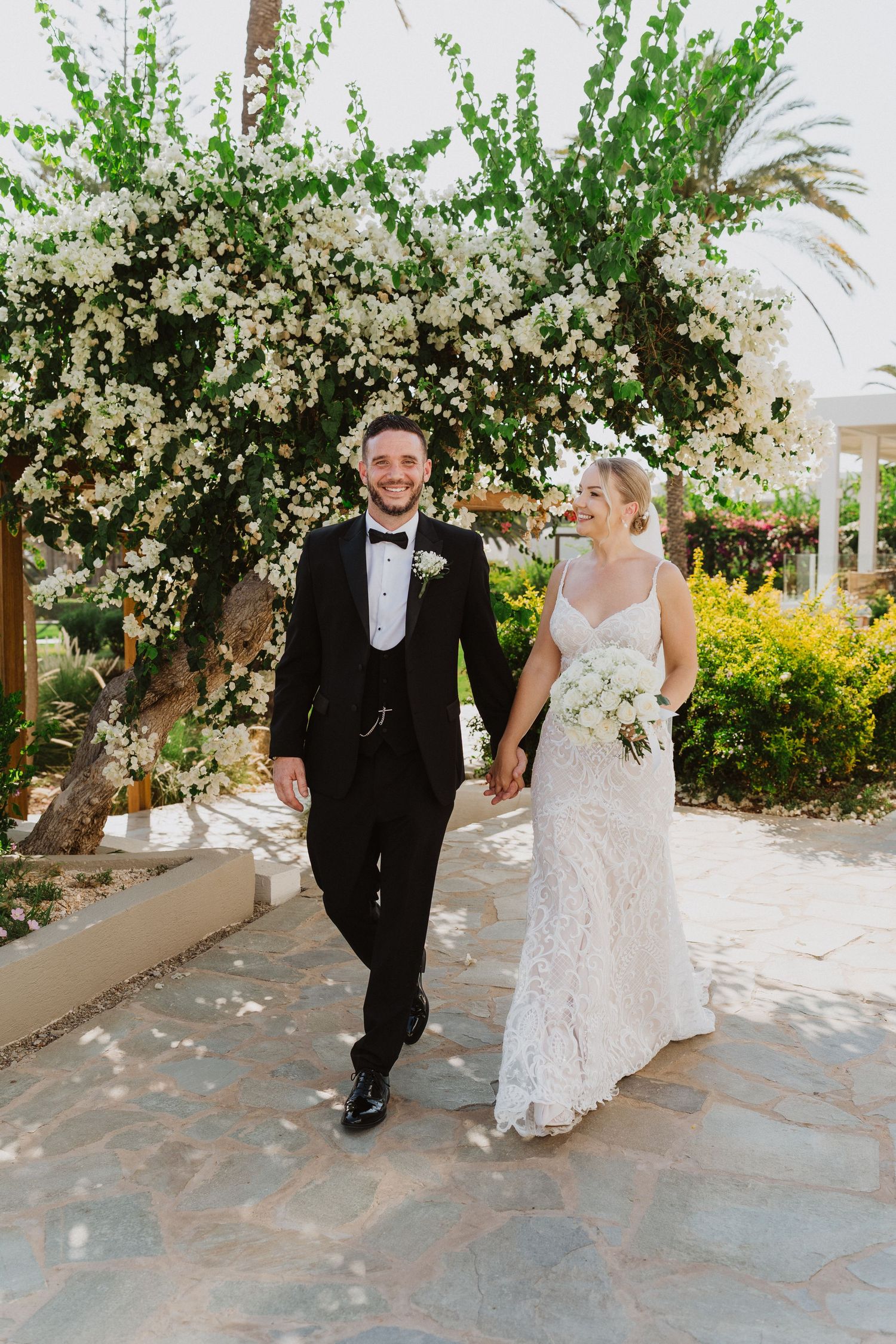 A couple in formal wedding attire walking together under blooming white floral archways in a Mediterranean garden setting.