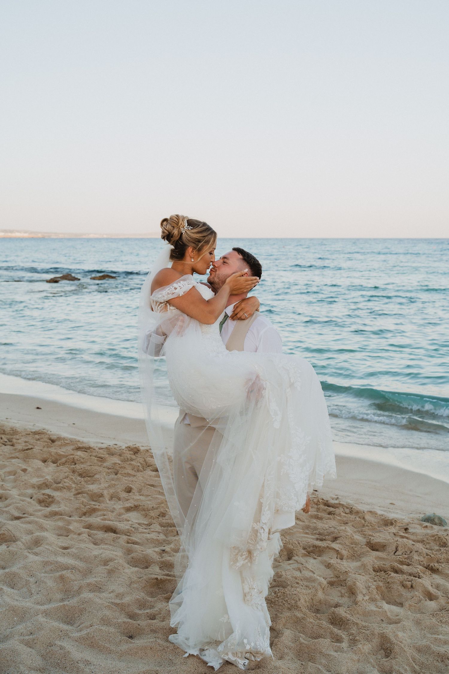 A romantic beach wedding couple embraces on the sandy shore at sunset with ocean waves in the background.