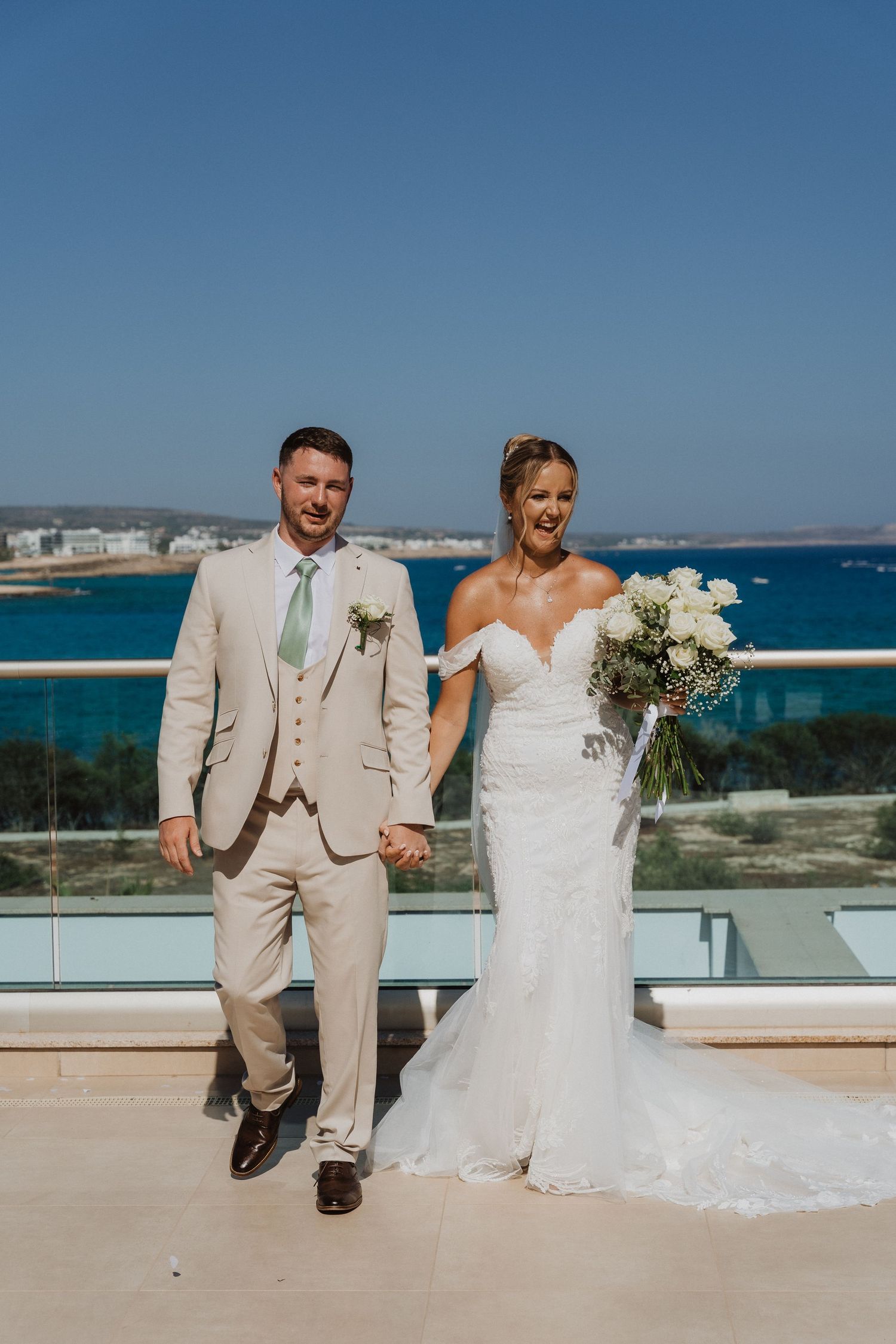 A wedding couple walks along a beachfront terrace with stunning blue ocean views in the background.
