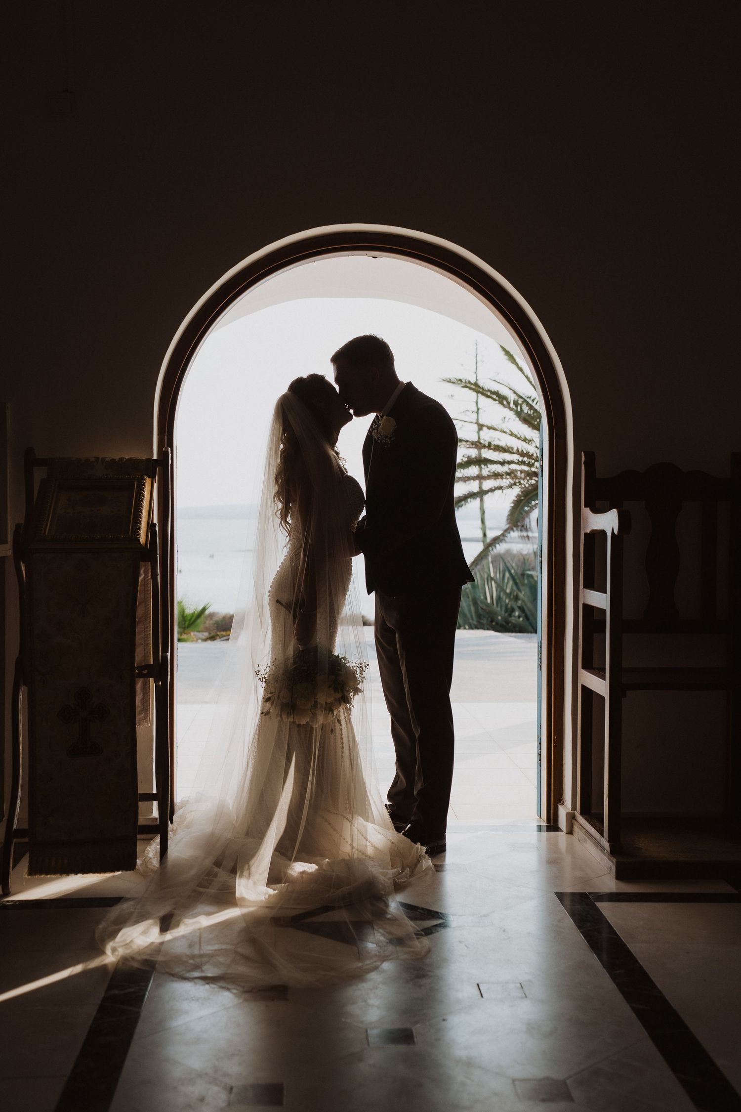 Romantic silhouette of bride and groom kissing in an arched church doorway with ocean view behind.
