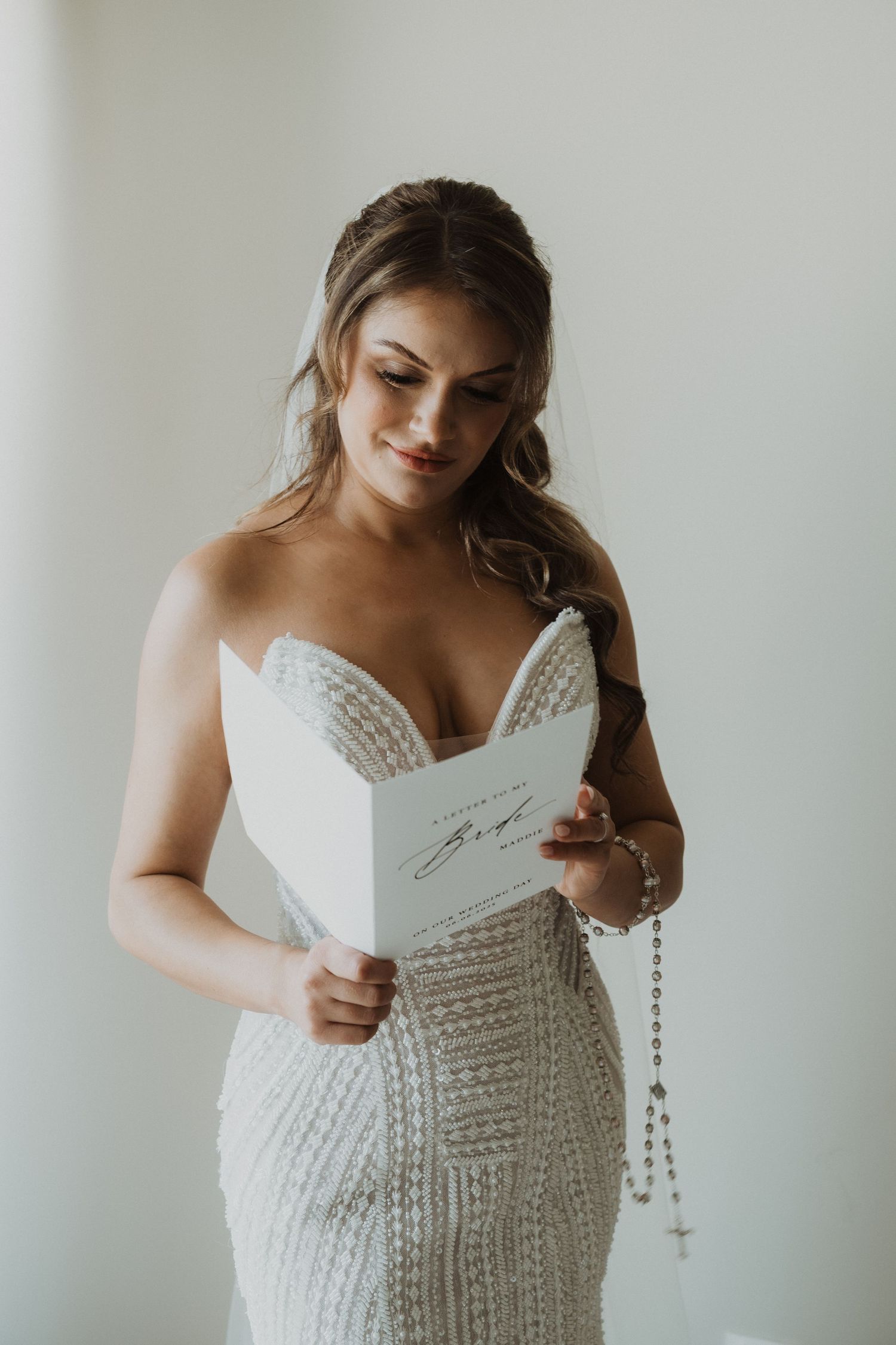 Beautiful bride in strapless beaded gown reading letter while holding rosary beads before ceremony.