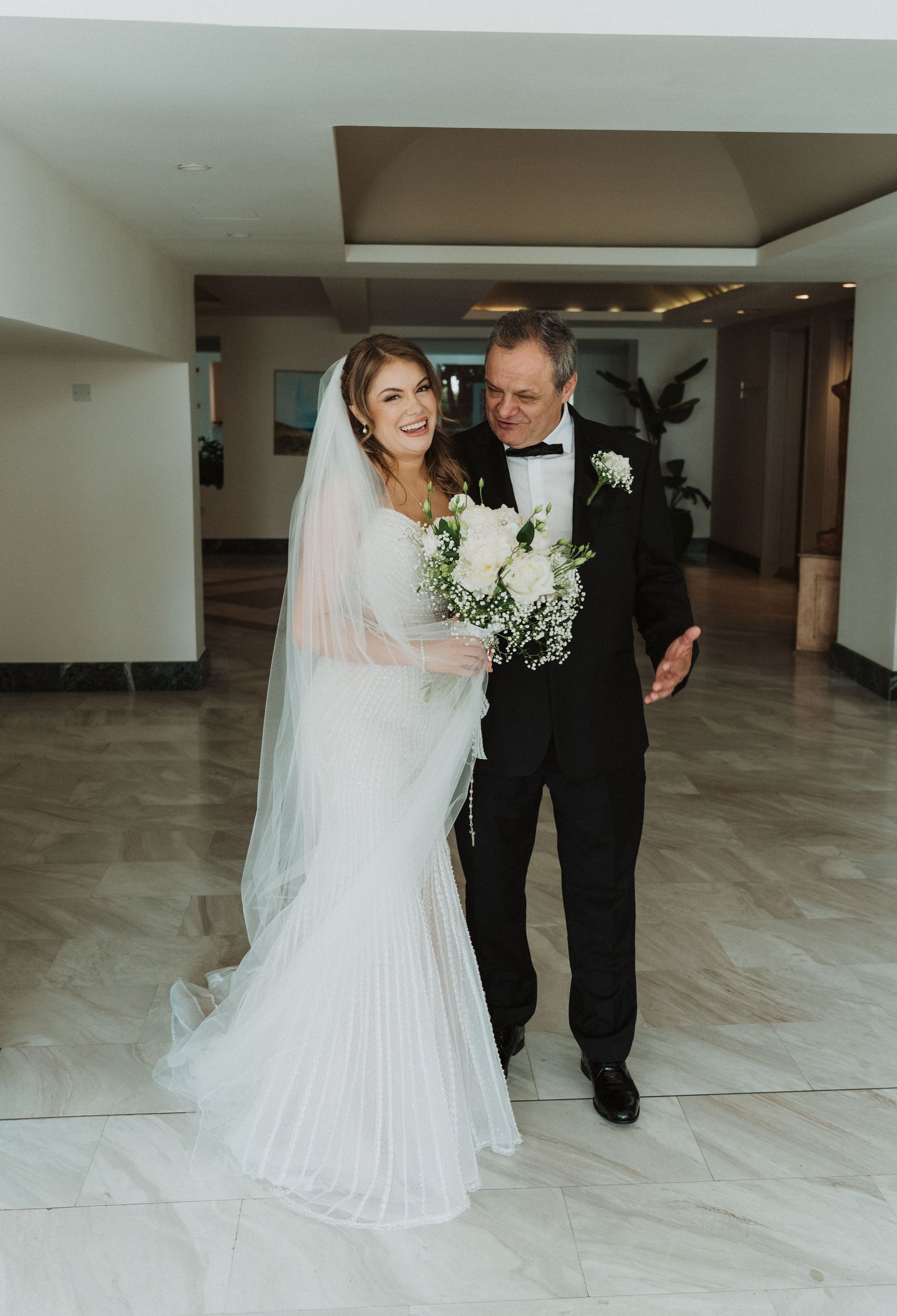 Radiant bride in mermaid wedding gown with veil walks with escort through elegant marble lobby.