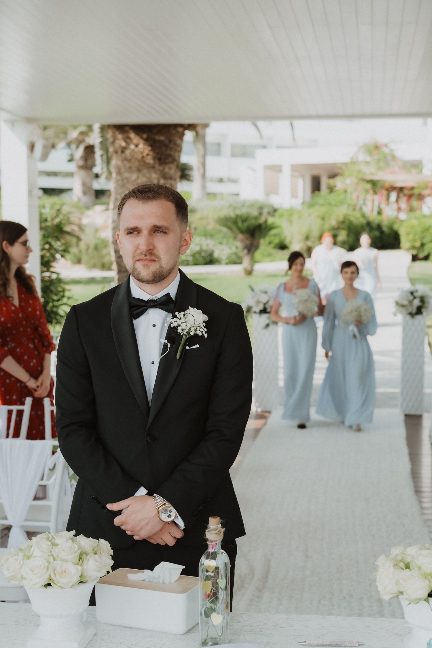 Groom in black tuxedo awaiting bride at altar as bridesmaids in blue dresses walk down aisle.
