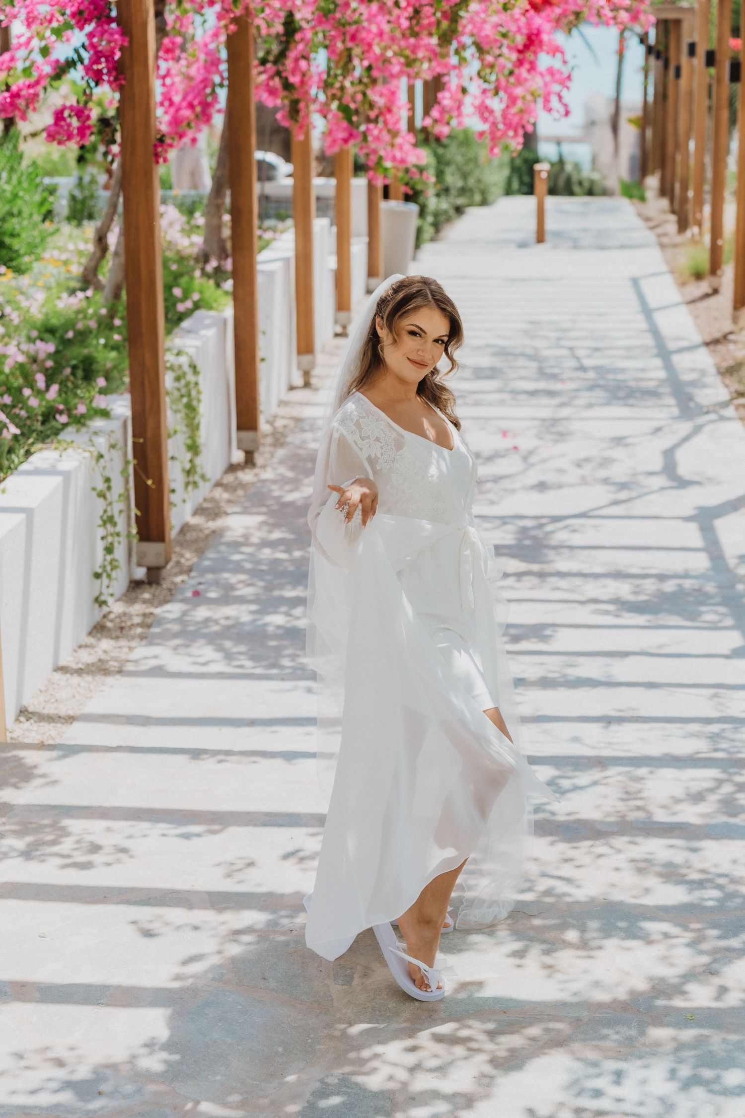 Beautiful bride in white dress walking under pink bougainvillea pergola.