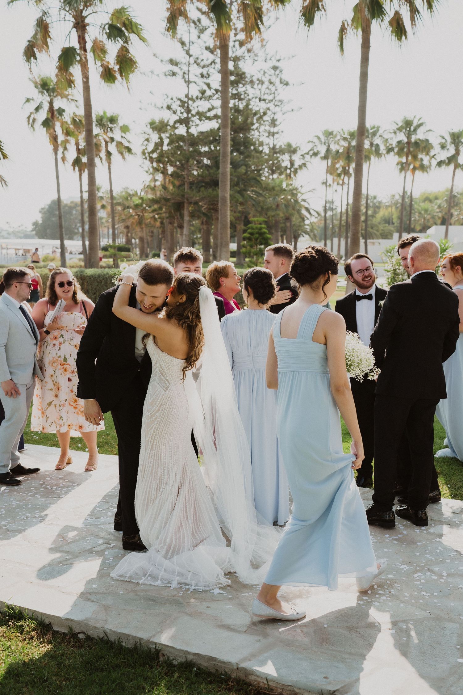 Wedding guests congratulate bride and groom outdoors surrounded by tall palm trees at elegant garden ceremony.