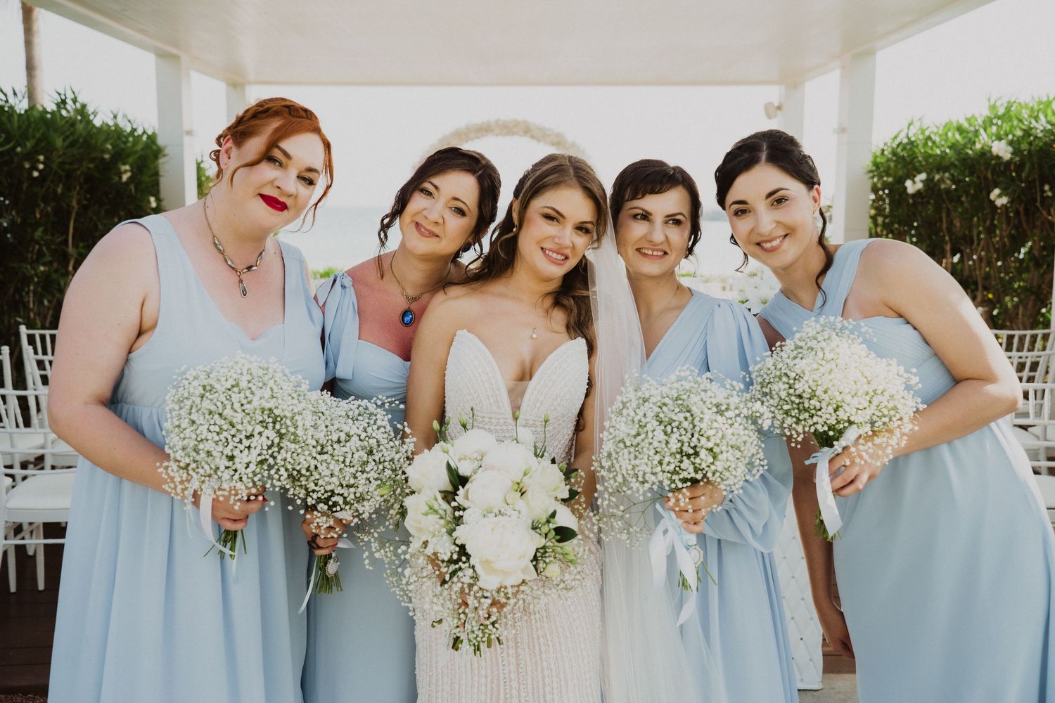 Bride in white gown with bridesmaids in light blue dresses holding white floral bouquets at an outdoor wedding ceremony.