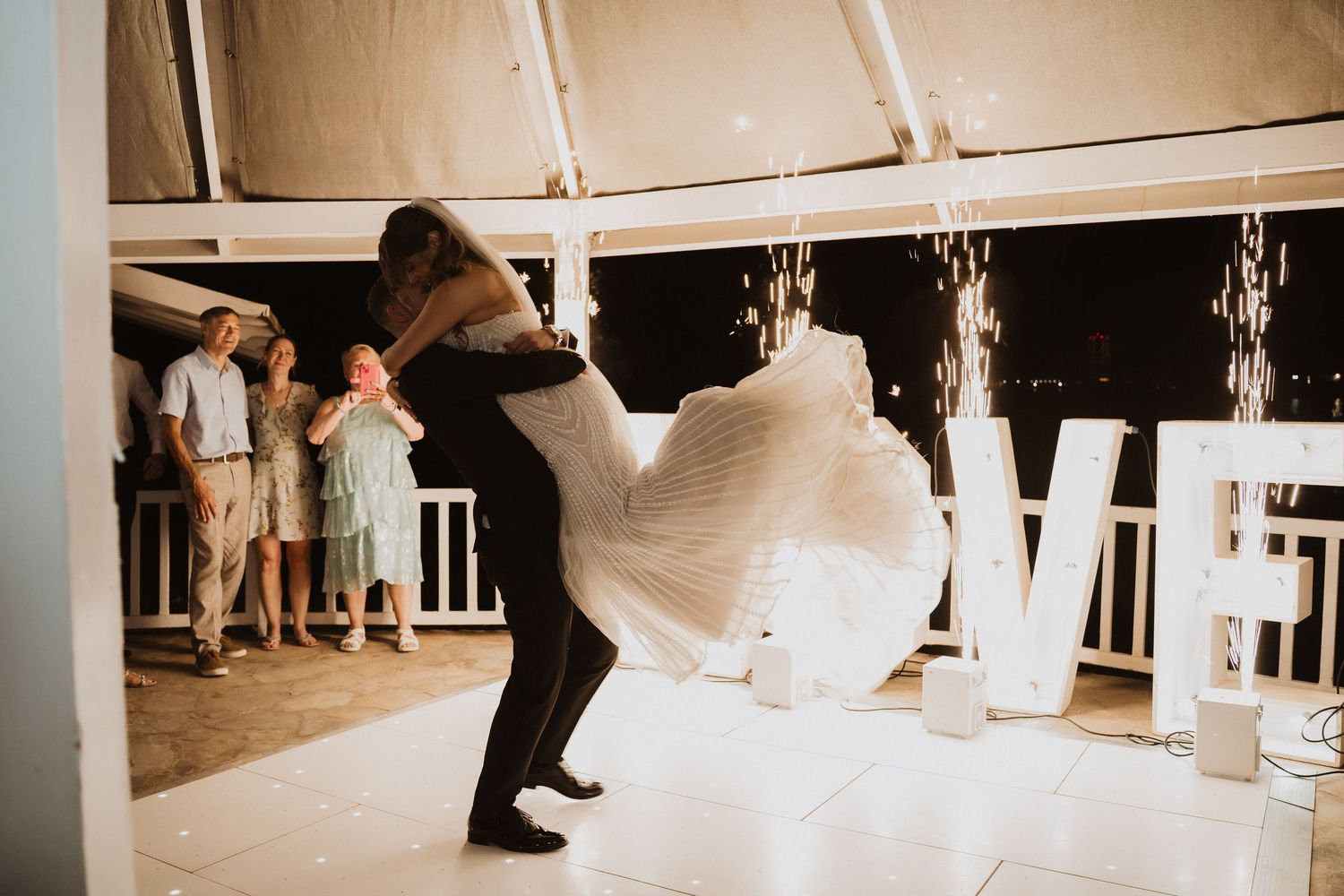 Groom lifts bride during first dance on illuminated dance floor with LOVE letters and sparklers nearby.