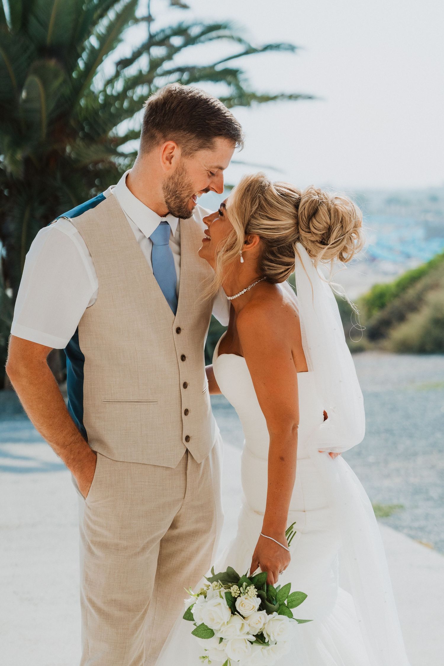 Bride and groom share a romantic moment, touching foreheads, as she holds a white bouquet at an outdoor wedding.