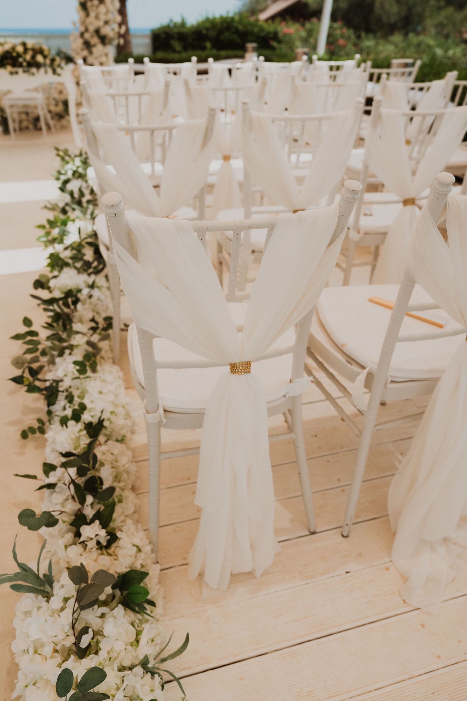 Elegant white chiavari chairs with draped fabric sashes and floral aisle decorations at an outdoor wedding ceremony.