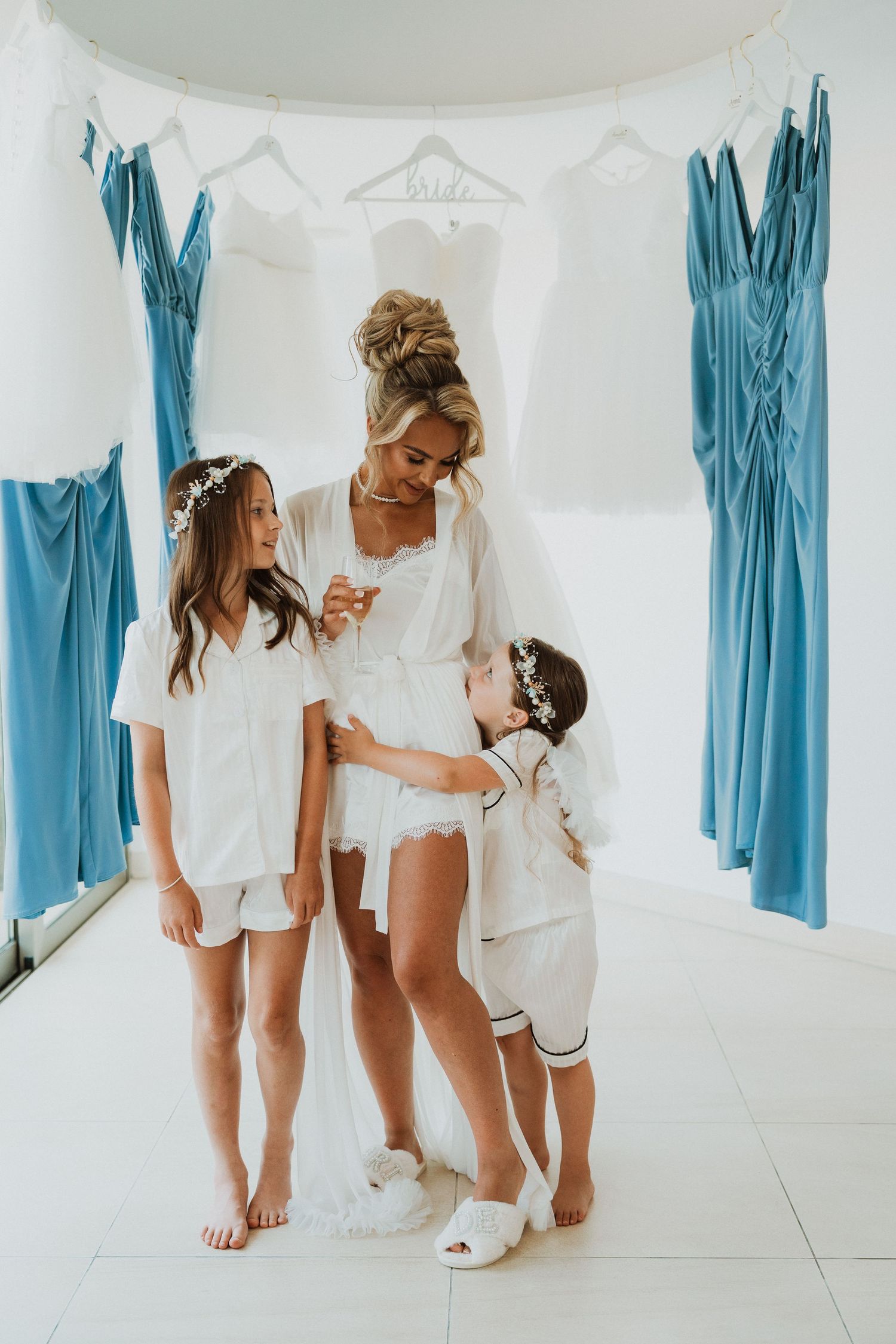 Bride in white robe with two flower girls in pajamas surrounded by blue bridesmaid dresses on wedding morning.
