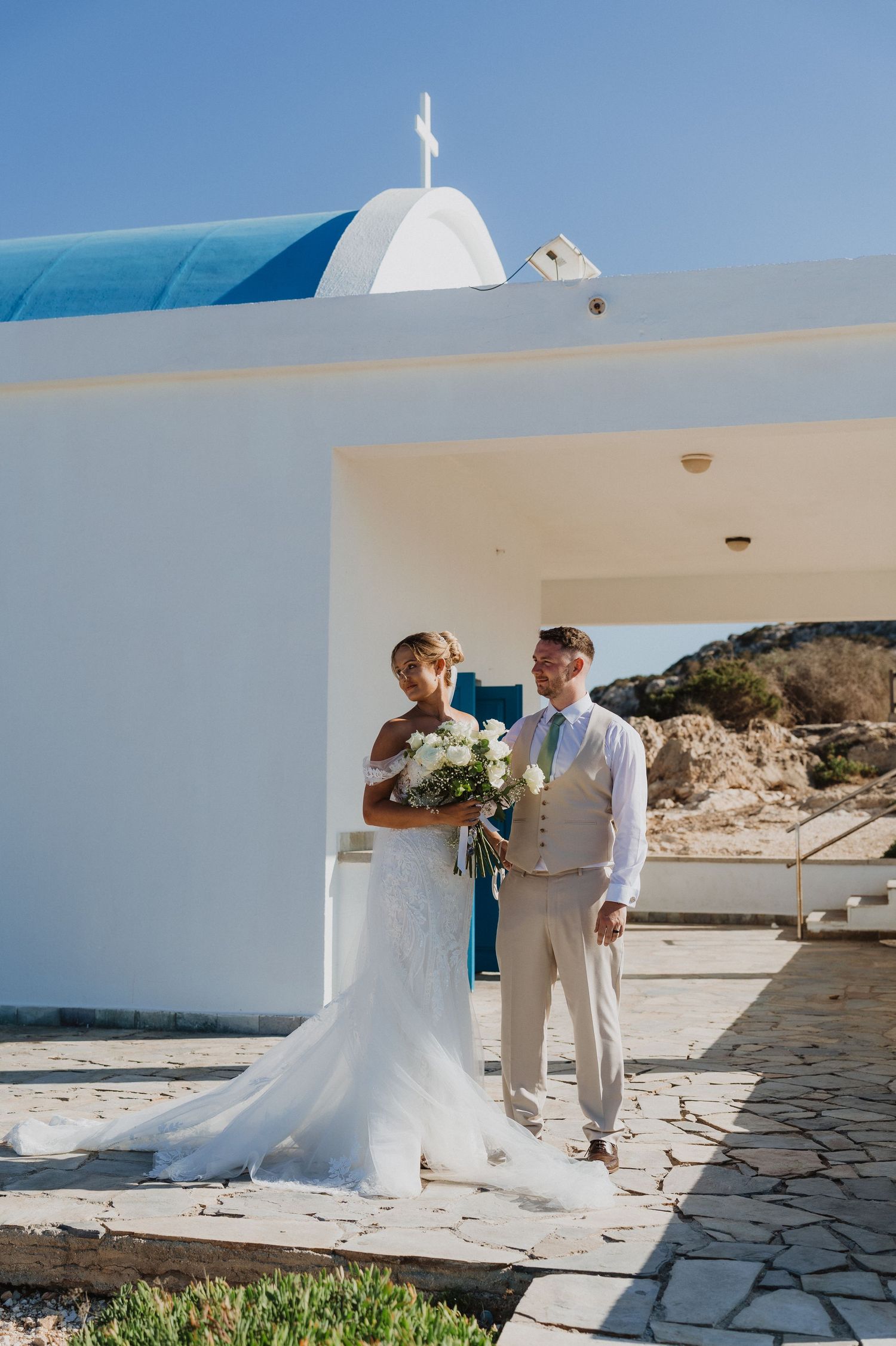 A couple stands outside a white Mediterranean church with blue dome in Cyprus on their wedding day.
