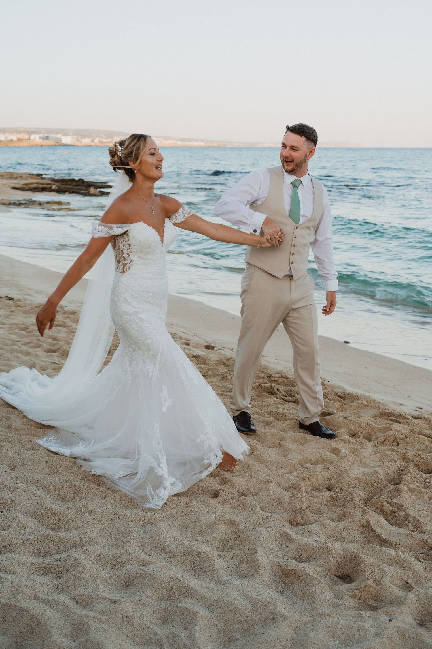 A couple in wedding attire playfully dances and spins together on a sandy beach at sunset with ocean waves in background.
