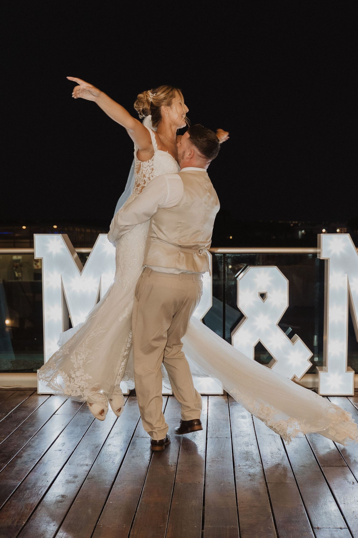 A couple in wedding attire dances together on a wooden deck at night with illuminated letters in the background.