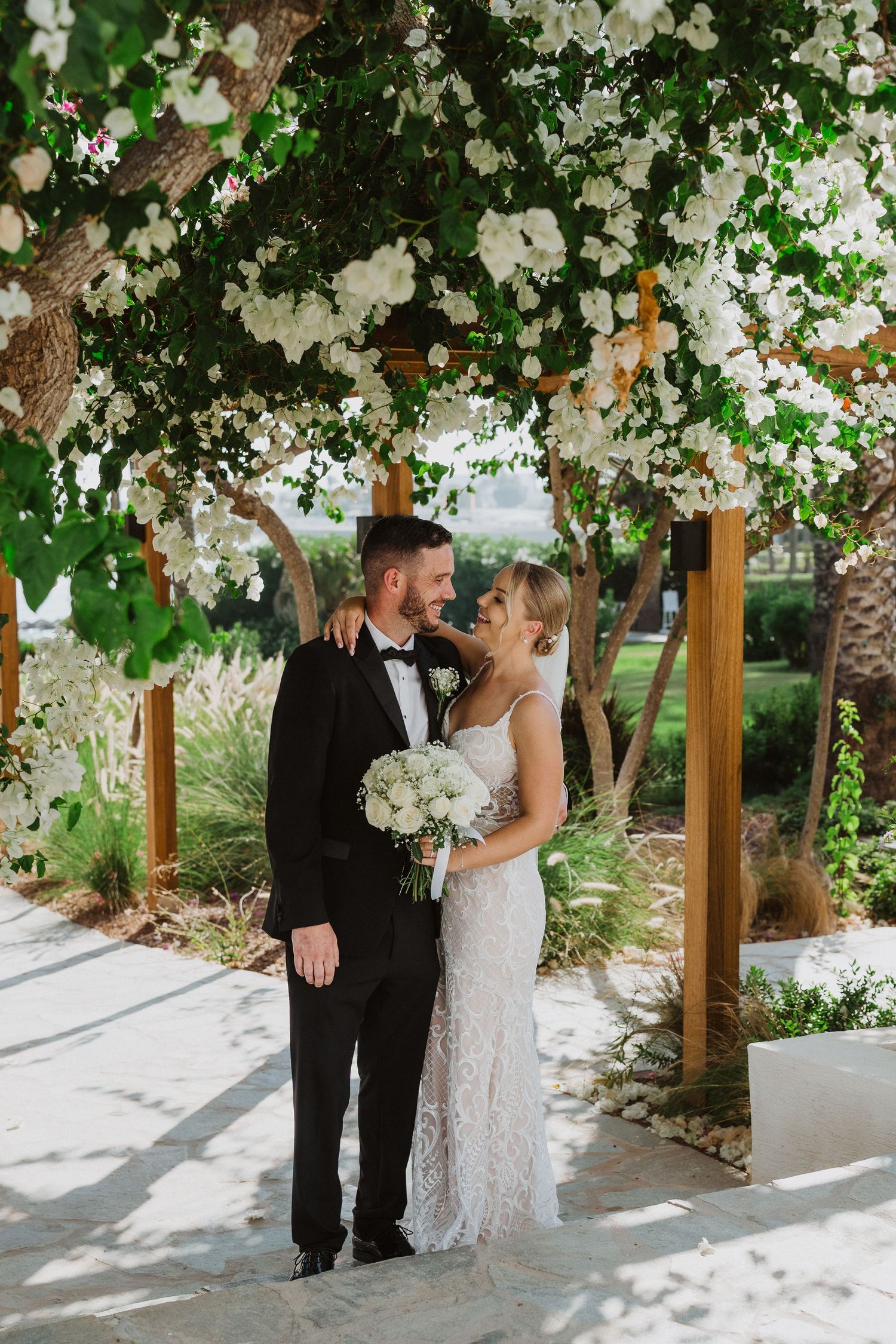 Newlyweds share a romantic moment under a floral-covered pergola in a garden setting.
