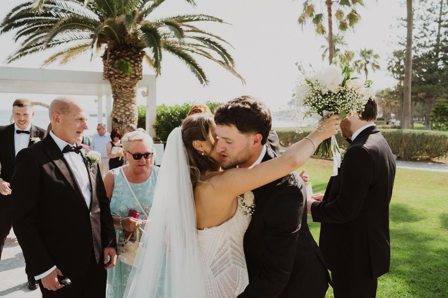 Bride and groom share their first kiss during an outdoor wedding ceremony surrounded by guests and palm trees.