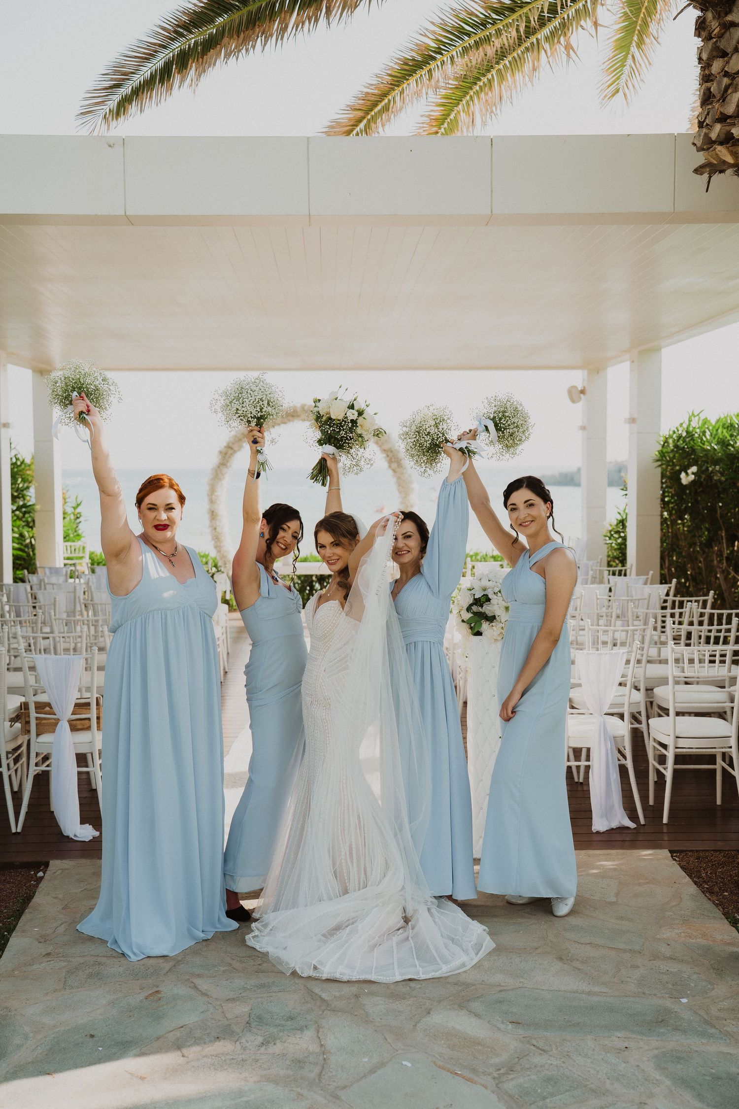 Bride poses with four bridesmaids in light blue gowns raising bouquets at outdoor wedding venue.