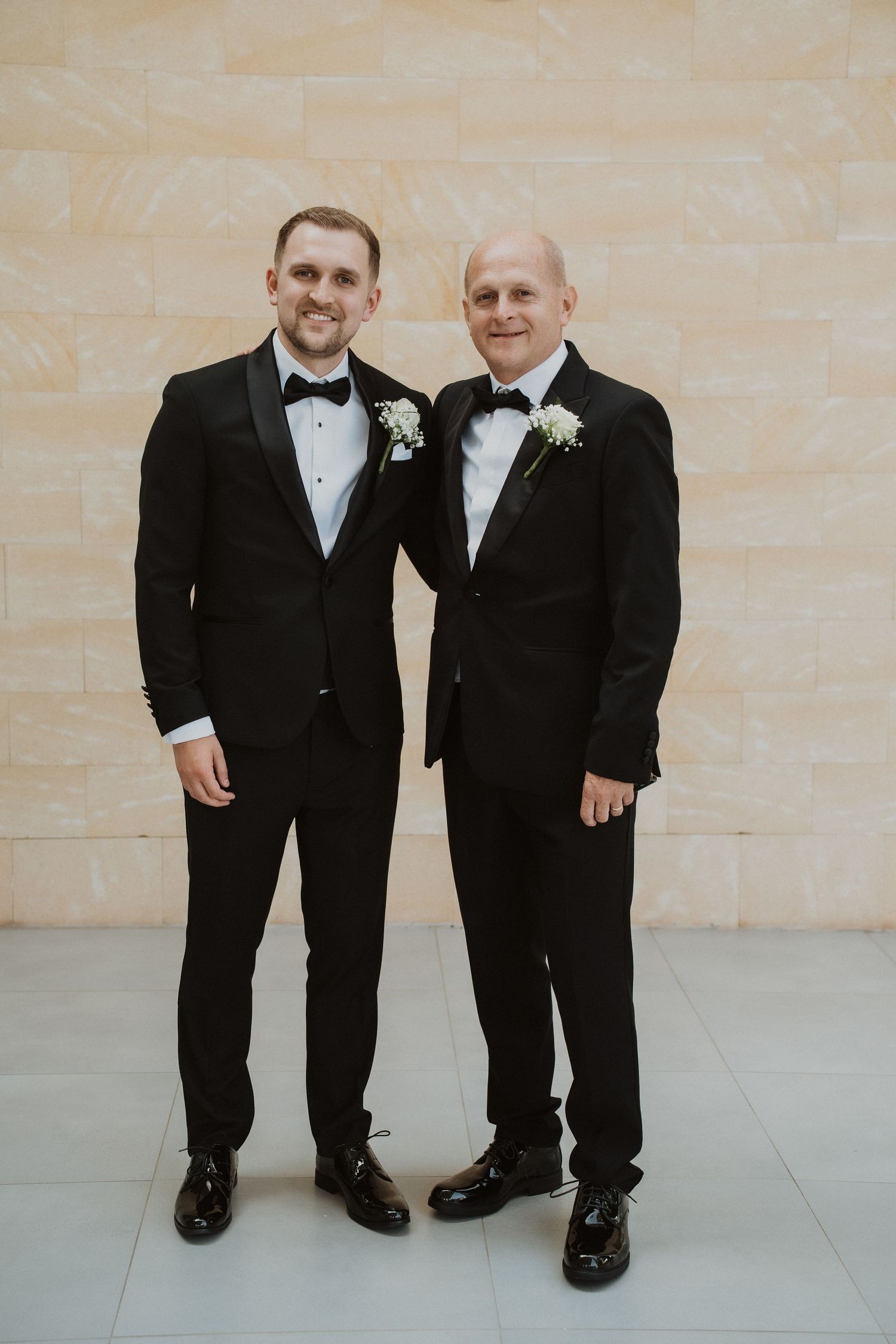 Two groomsmen in matching black tuxedos with boutonnieres posing together at wedding.