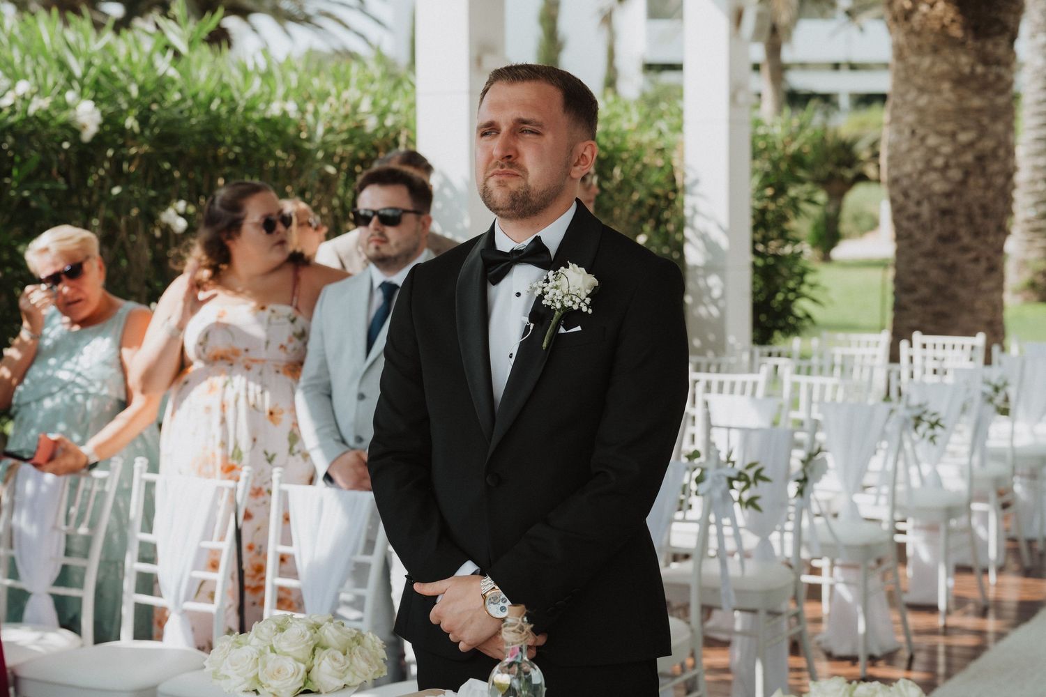 Groom in black tuxedo standing at the altar awaiting bride at outdoor wedding ceremony.