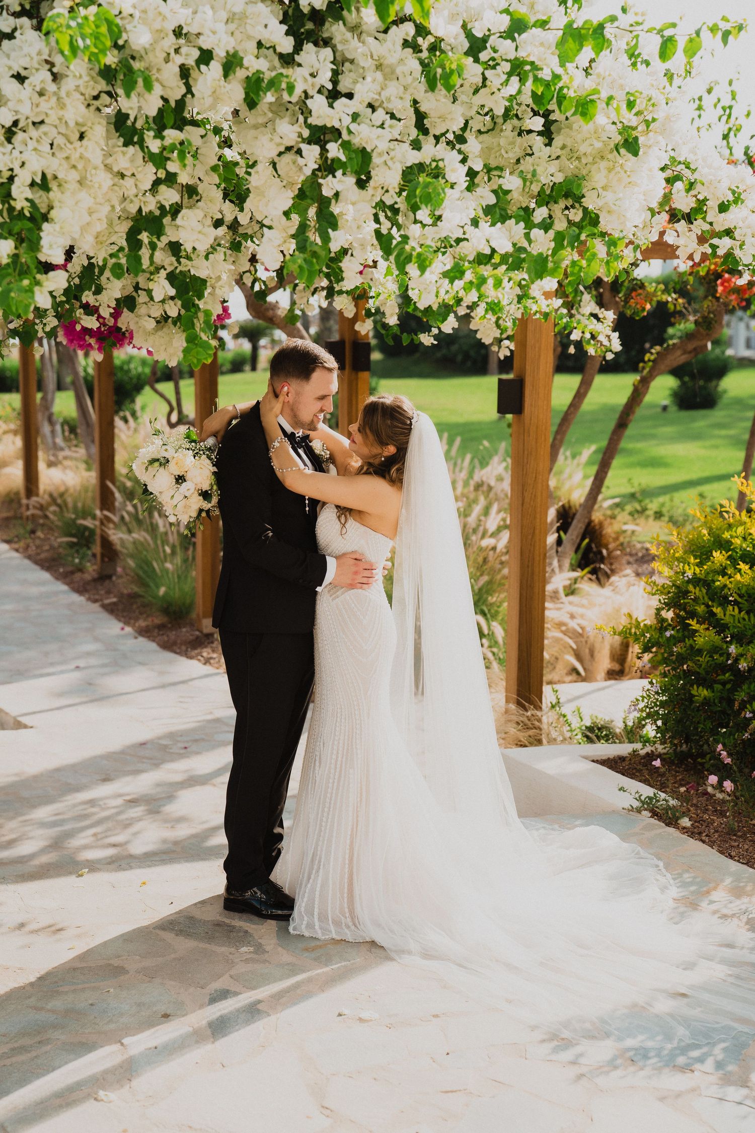 Newlyweds share a tender moment under a stunning white floral pergola surrounded by lush garden greenery.