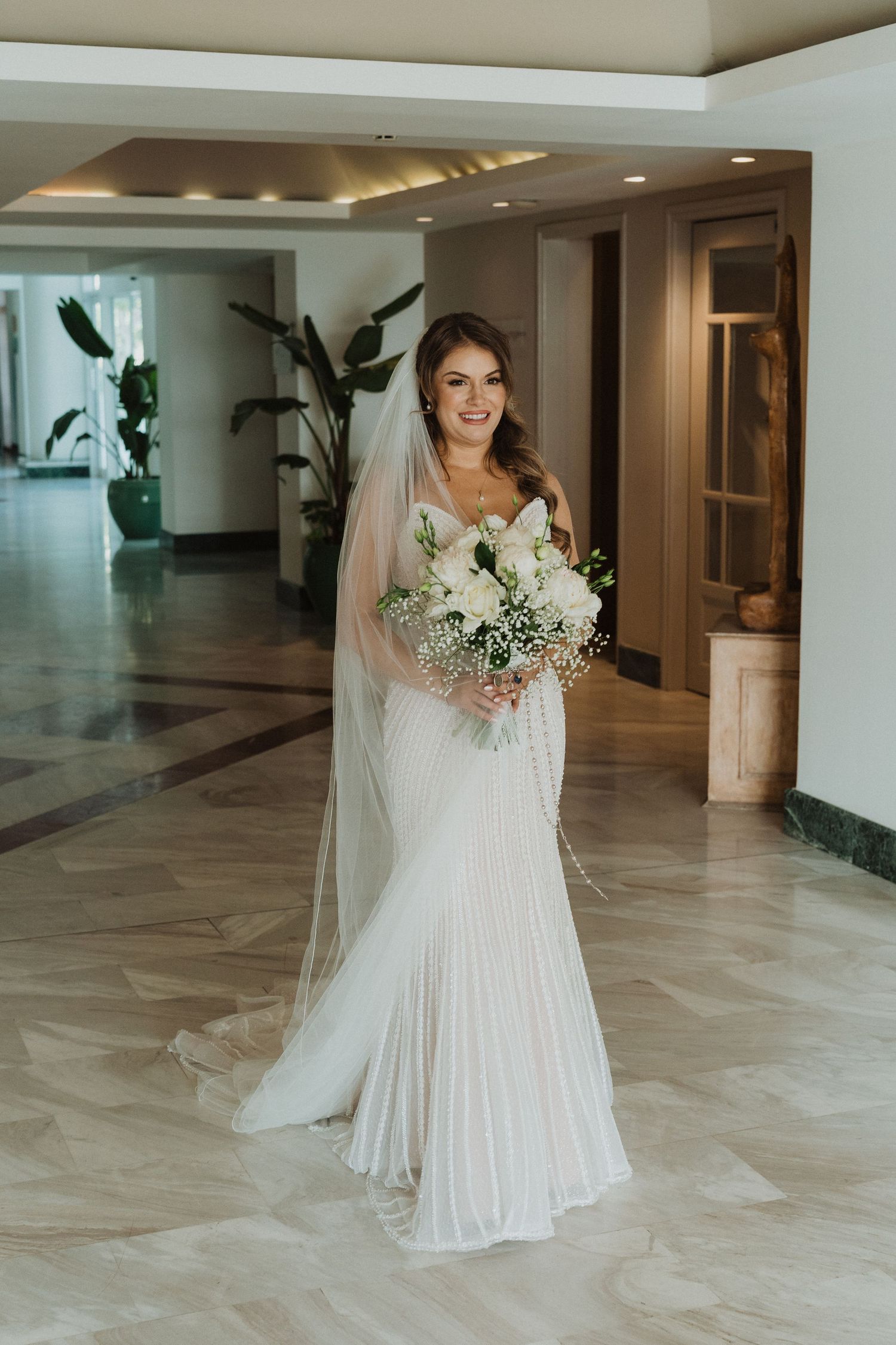 Smiling bride in a white pleated gown and long veil holds a white bouquet in an elegant hotel lobby.