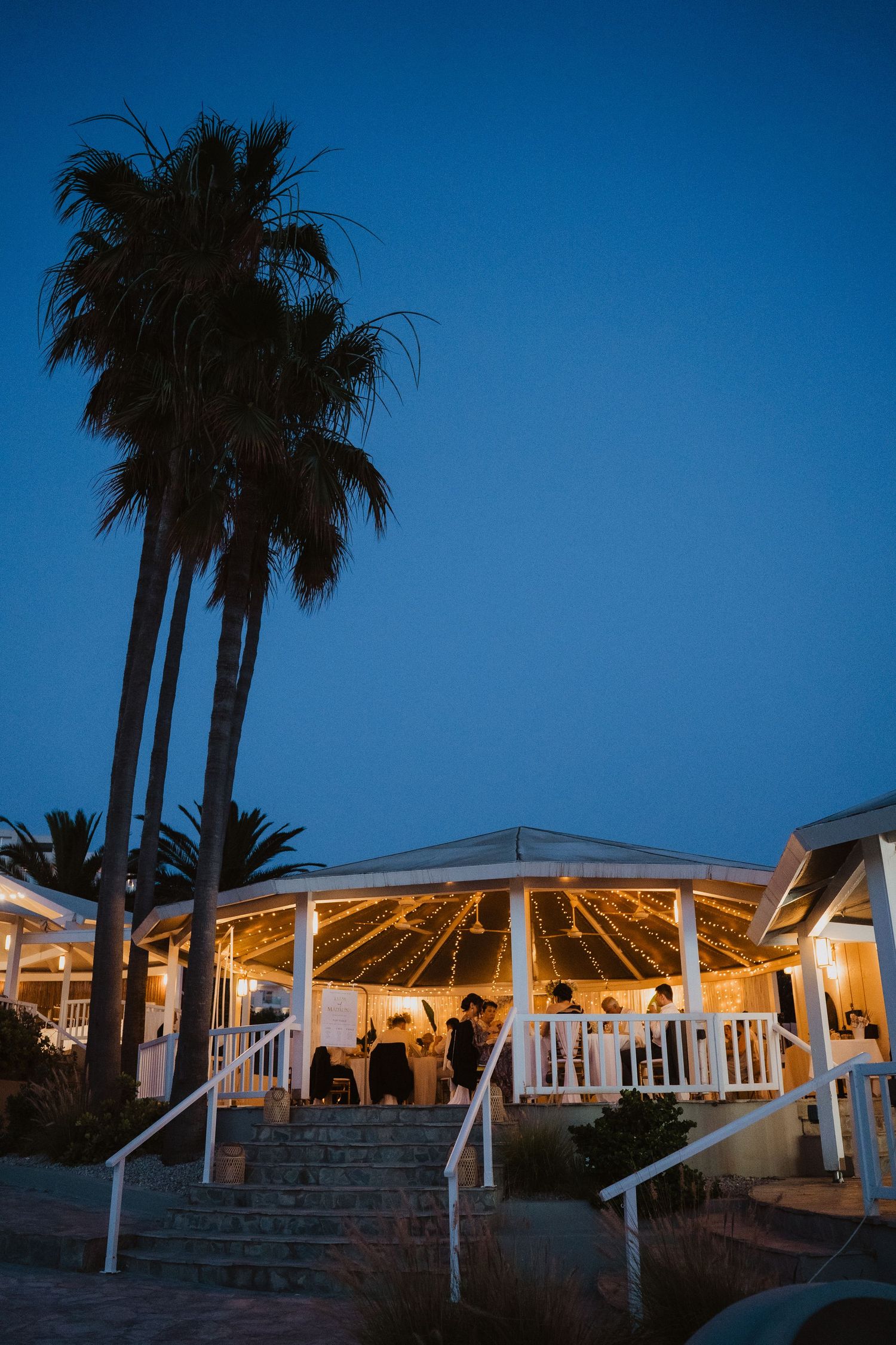 Illuminated outdoor wedding reception pavilion with string lights and palm trees silhouetted against deep blue evening sky.