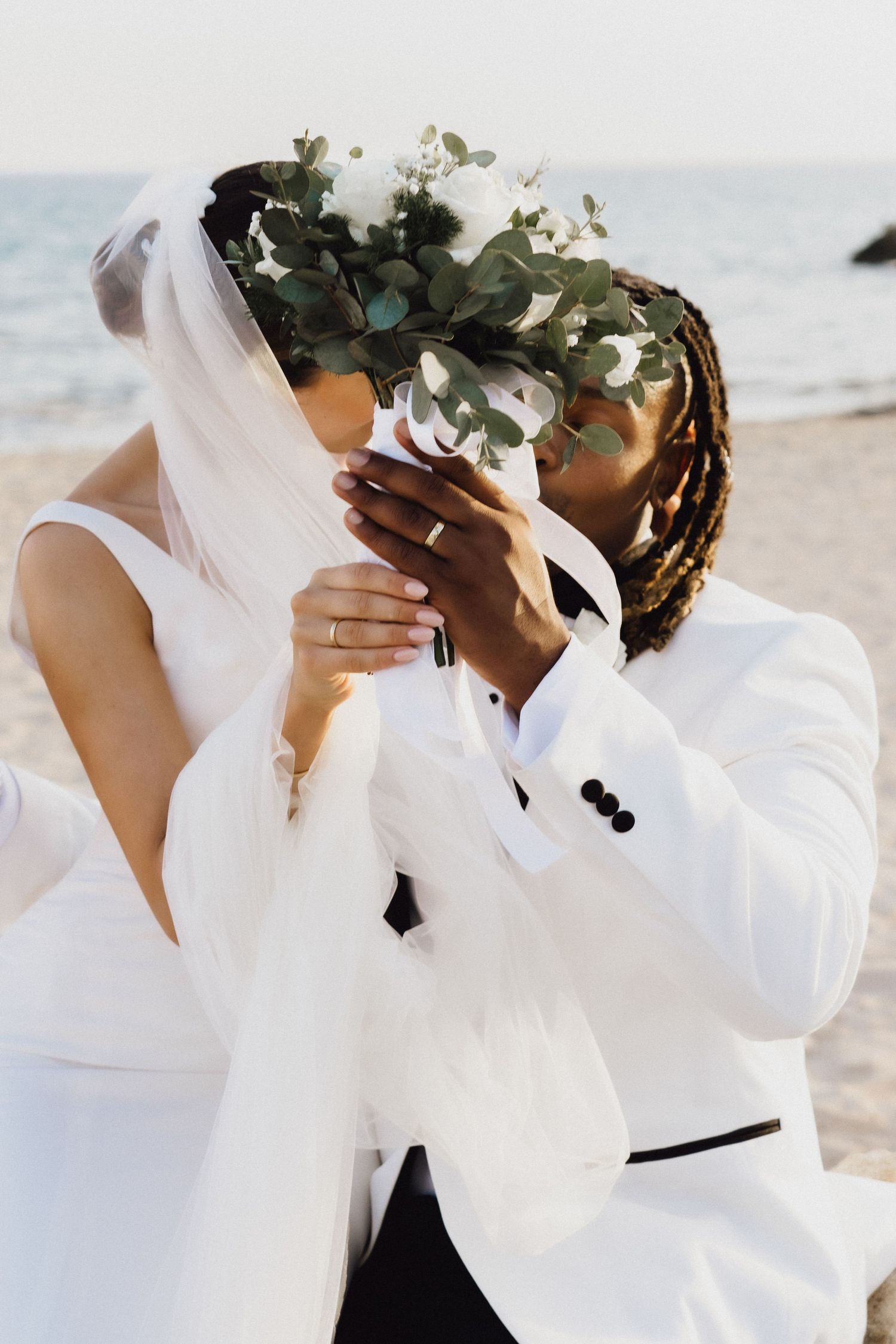 A couple shares an intimate moment behind a wedding bouquet featuring eucalyptus on a sandy beach.