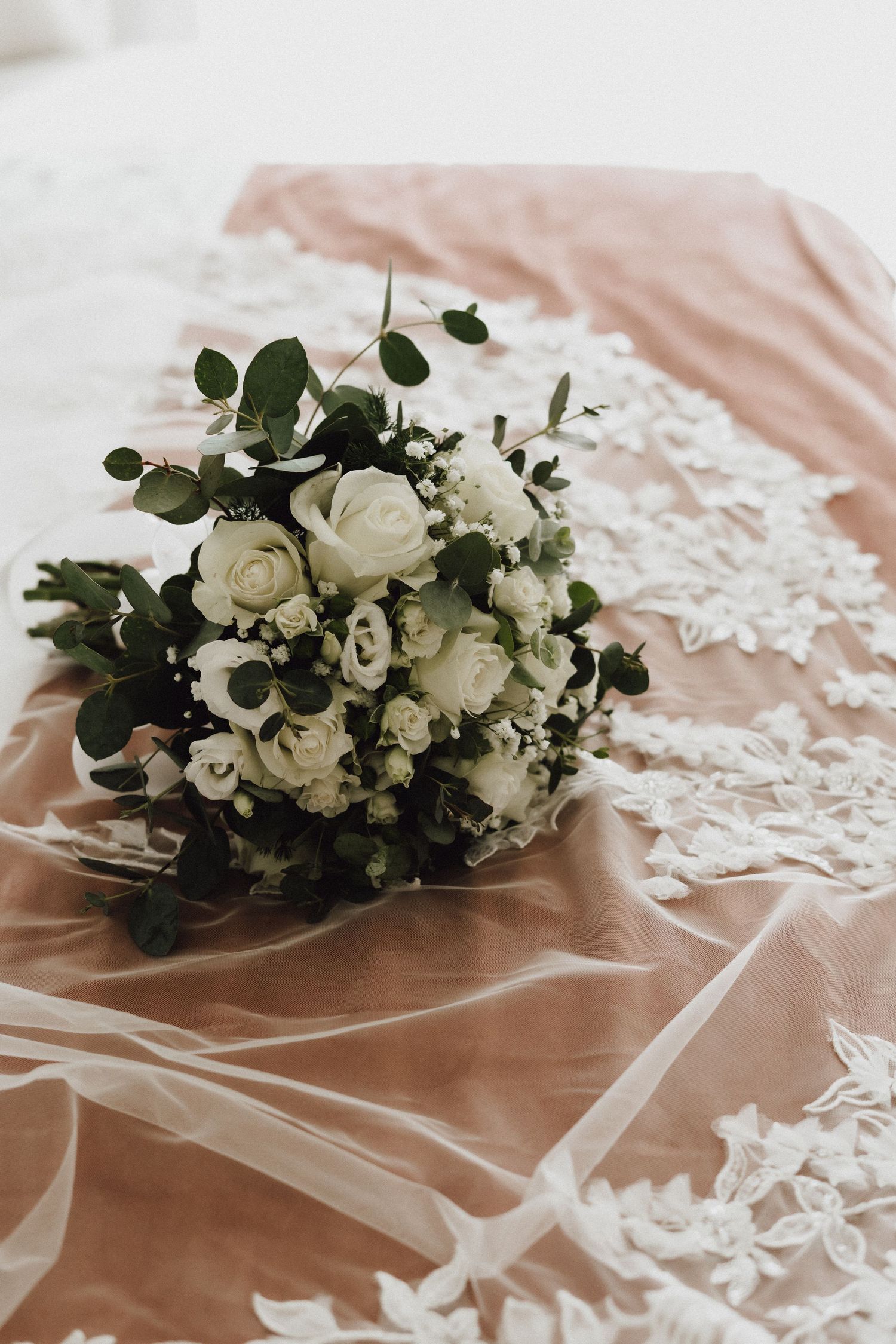 White rose bridal bouquet with eucalyptus leaves resting on pink silk and lace fabric.