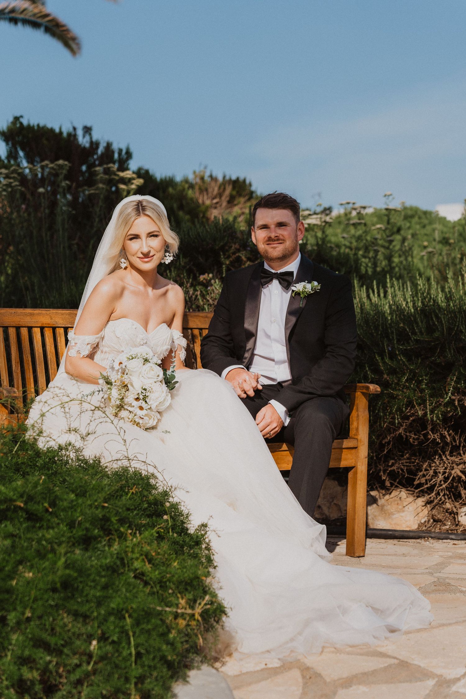 Newlyweds relaxing on wooden bench surrounded by coastal garden landscaping.