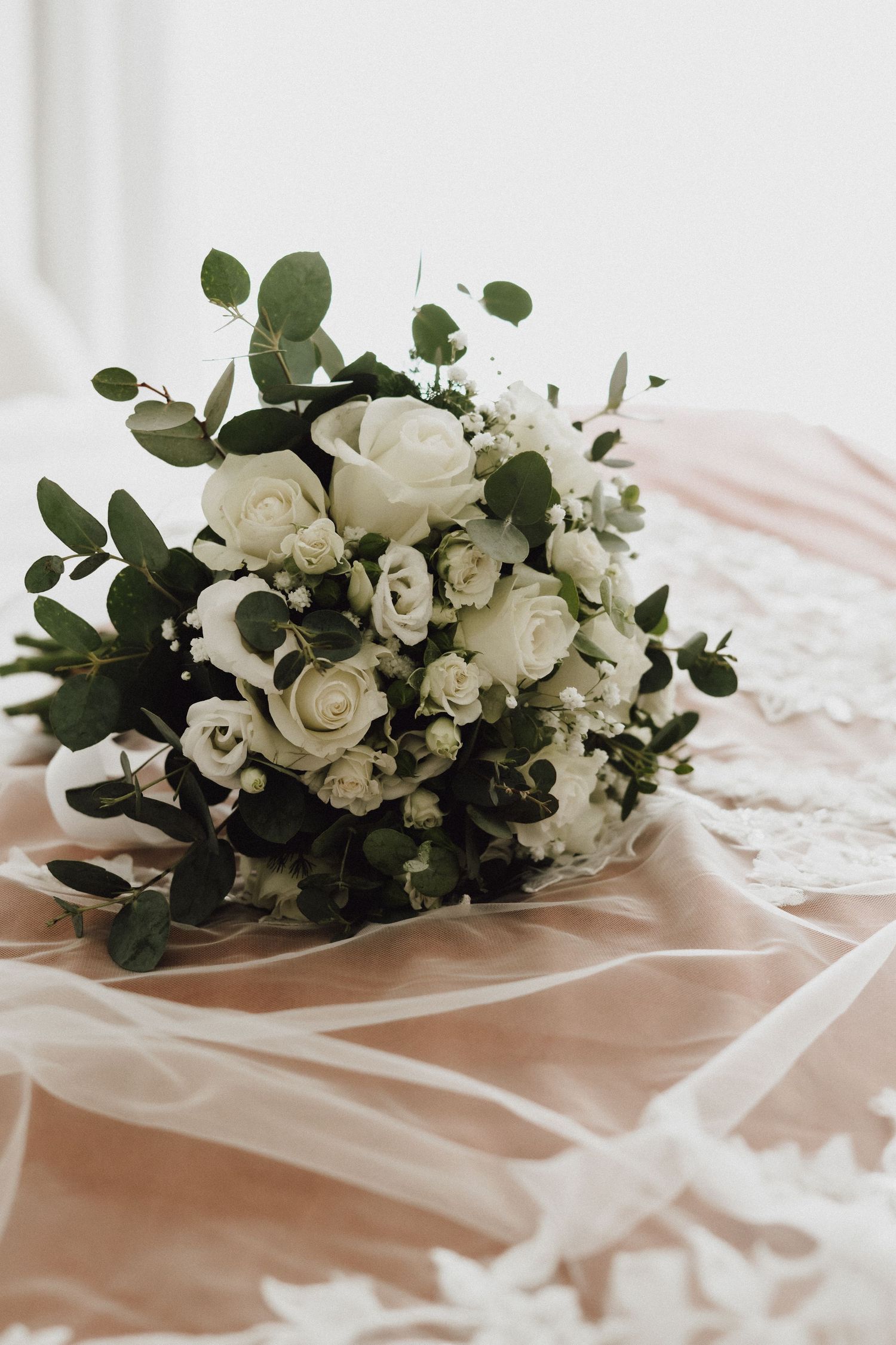 Classic white and green wedding bouquet featuring roses and eucalyptus on blush fabric.