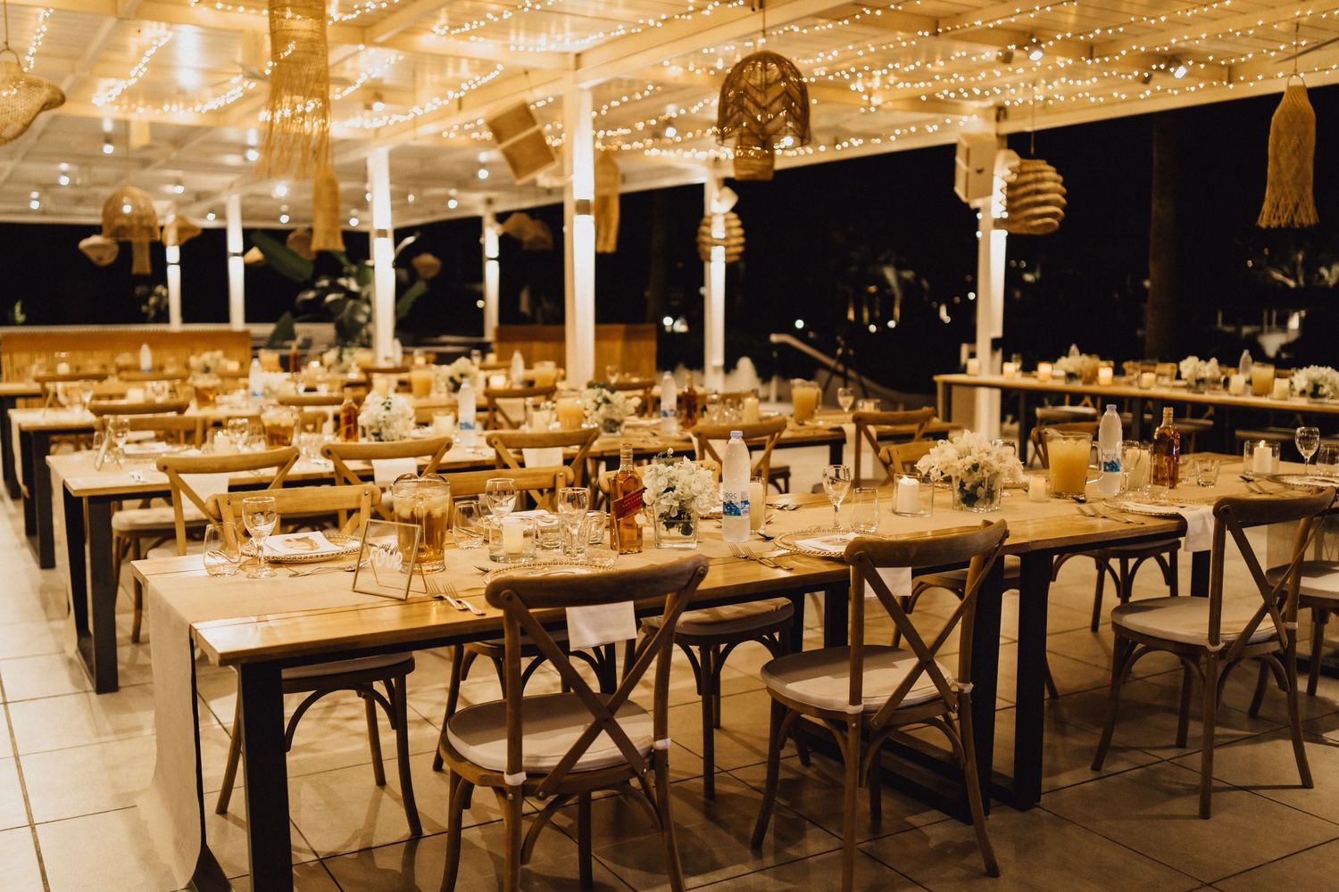 A rustic wedding reception setup with wooden cross-back chairs and long dining tables.