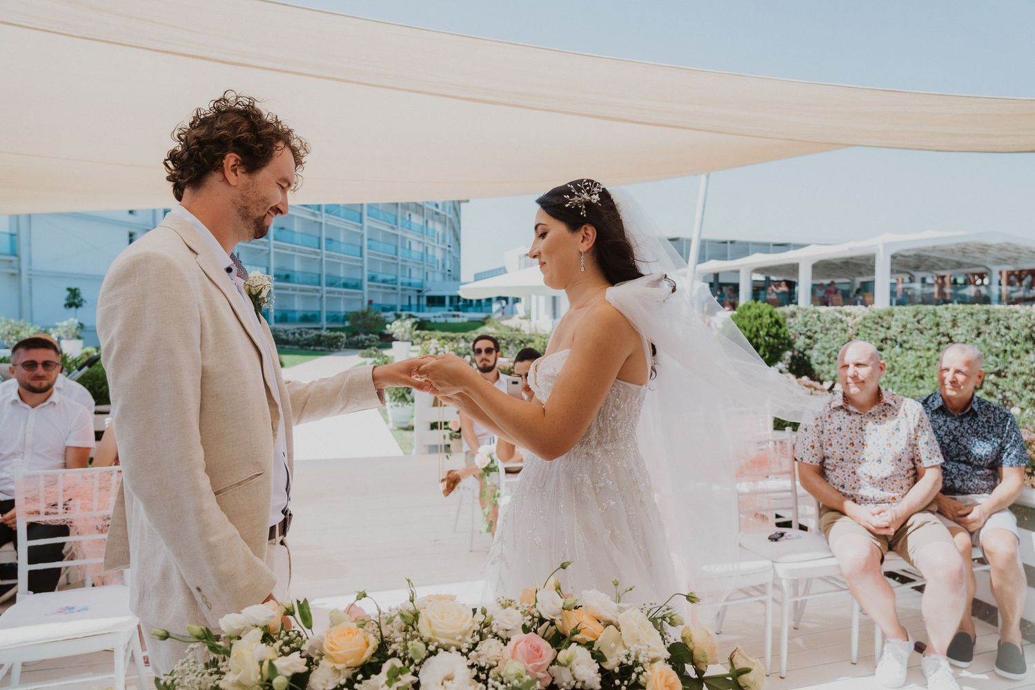 Couple exchanging vows during outdoor ceremony with white chairs and floral decorations.