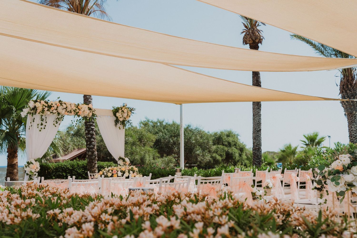 Outdoor wedding ceremony setup with white canopy sails, floral decorations and palm trees in a tropical garden setting.