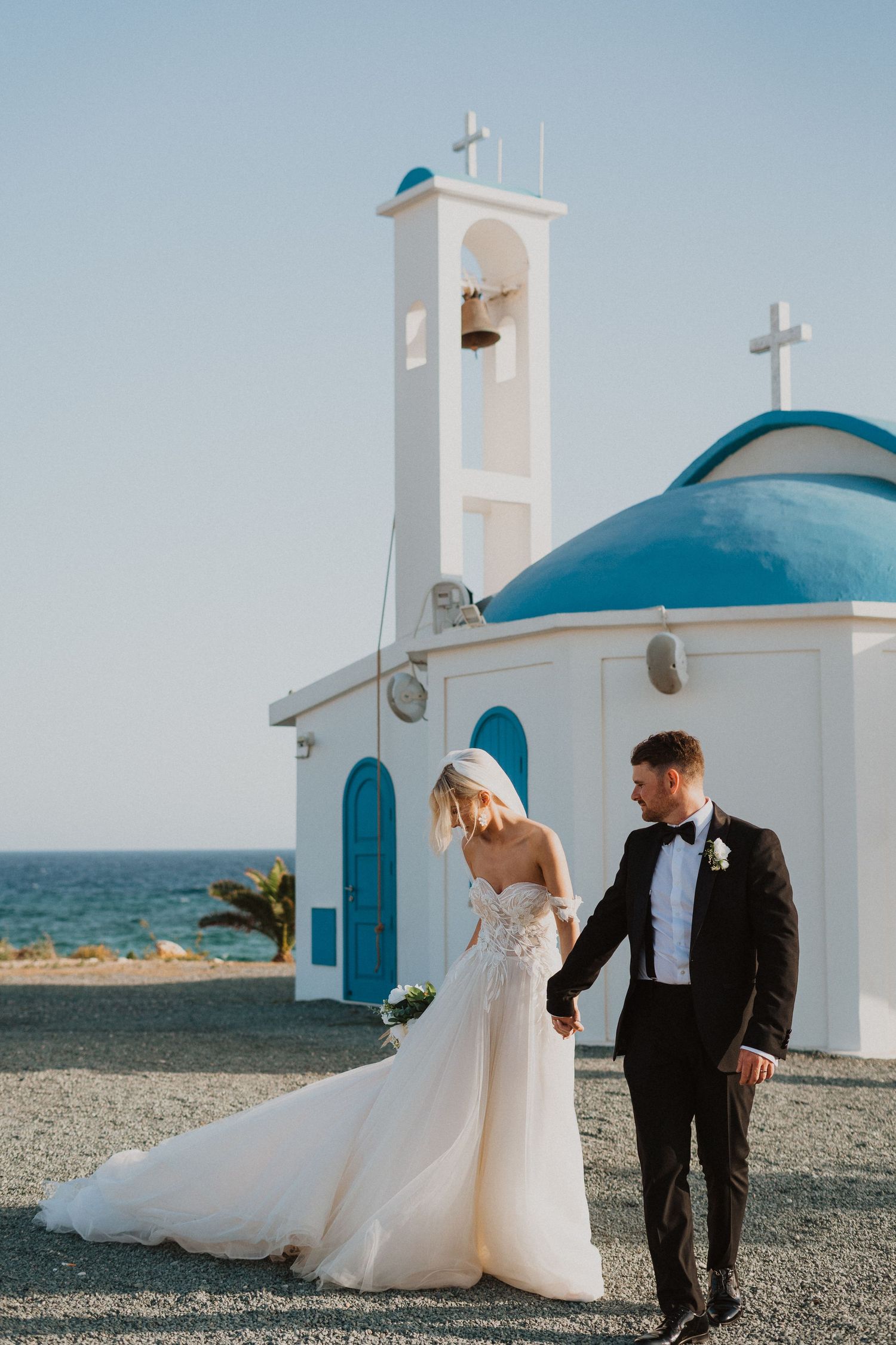 A wedding couple stands in front of a traditional Greek church with white walls and turquoise dome.