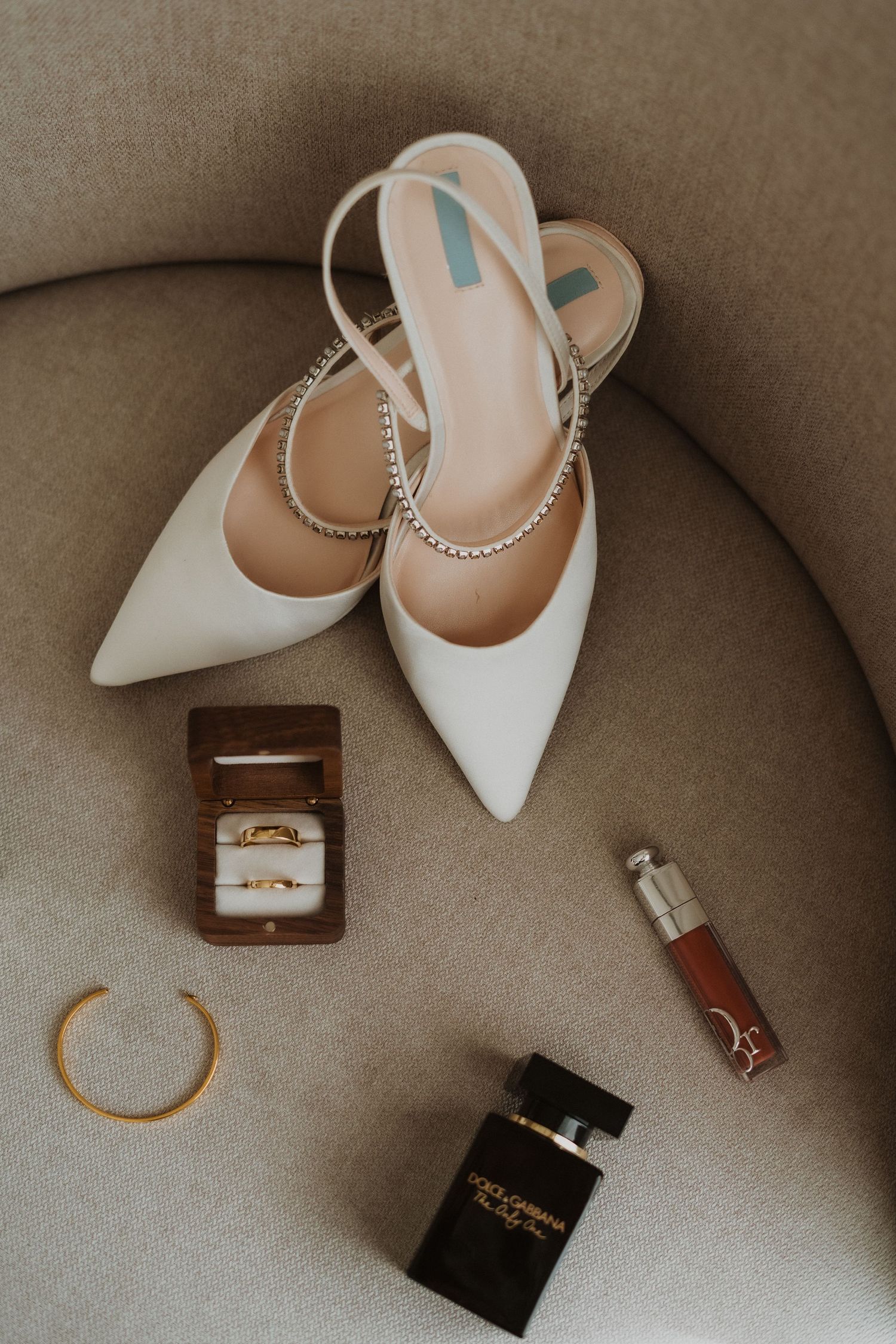 Elegant white bridal shoes with accessories and perfume displayed on a neutral chair.
