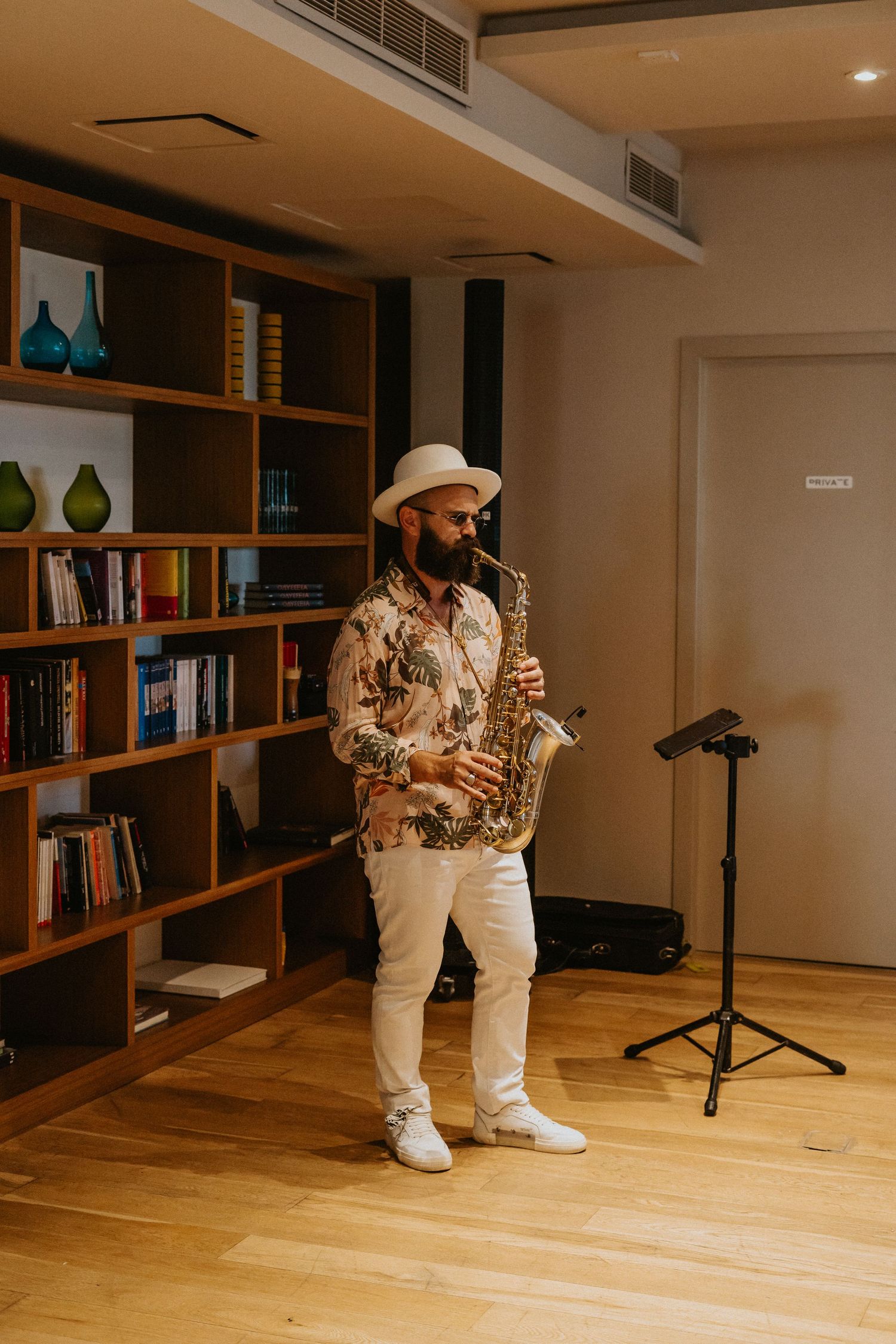 Musician in Hawaiian shirt and hat plays saxophone in front of wooden bookshelves in warm-lit indoor space.