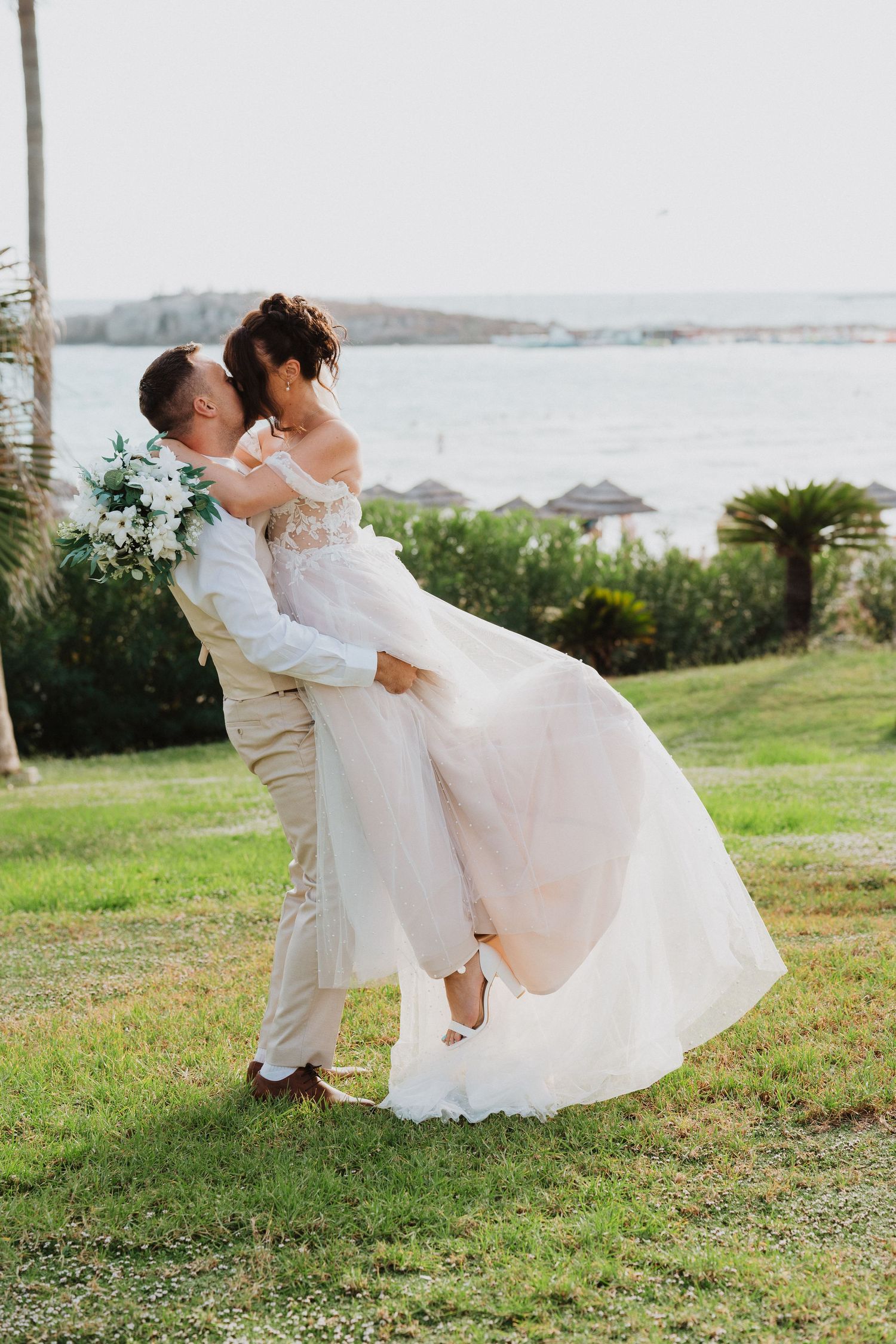 A romantic wedding couple embraces on a hilltop overlooking the ocean, with the bride wearing a flowing white gown.