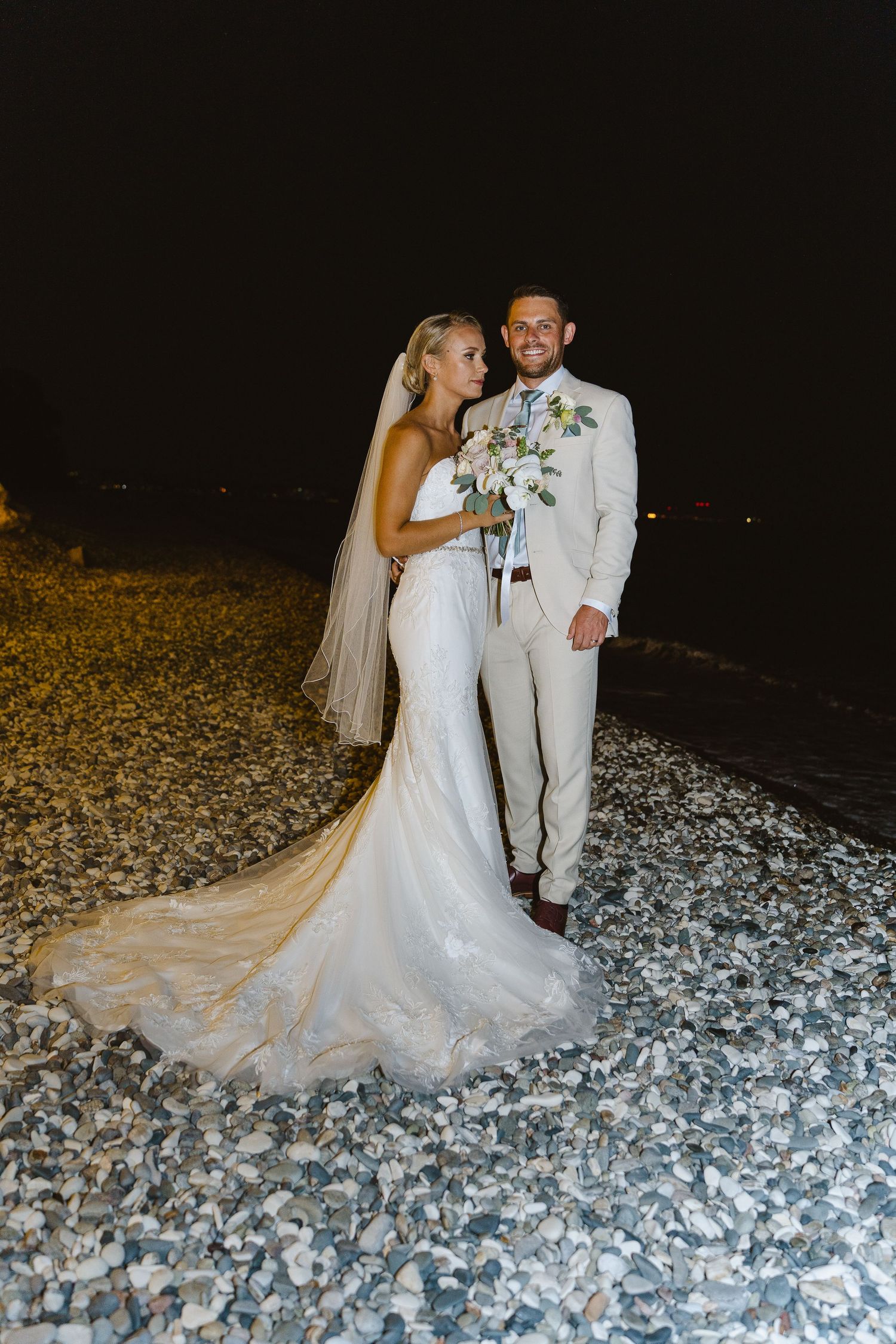 Wedding couple poses together on rocky beach at night with bride in flowing lace gown.