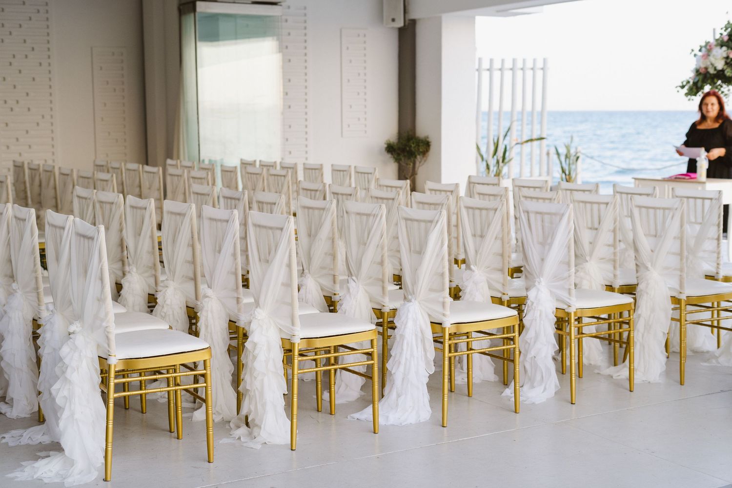 White fabric-draped chairs with gold accents line up for a beachside wedding ceremony.