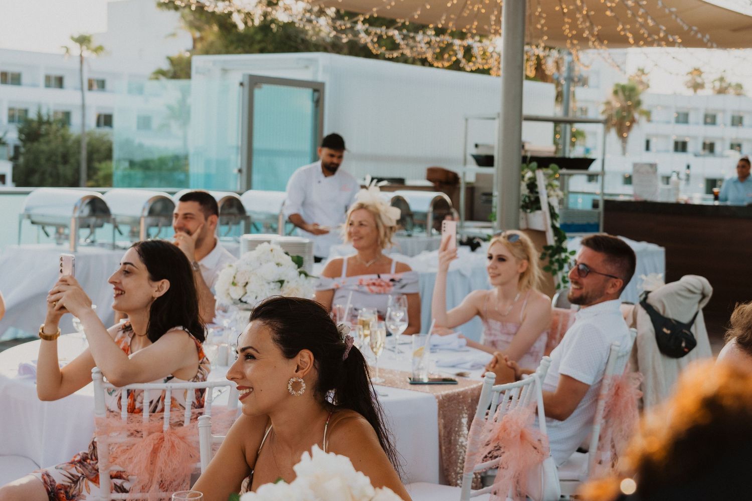 Evening wedding reception with guests enjoying drinks at decorated outdoor tables.
