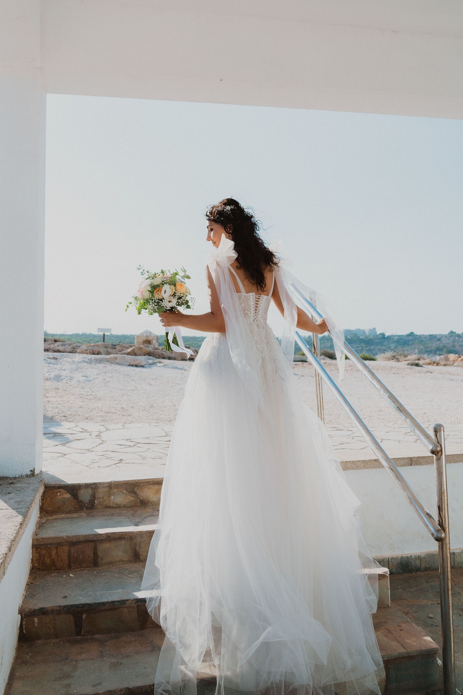 A bride in a flowing white gown holds a bouquet while standing on outdoor stairs against a bright backdrop.
