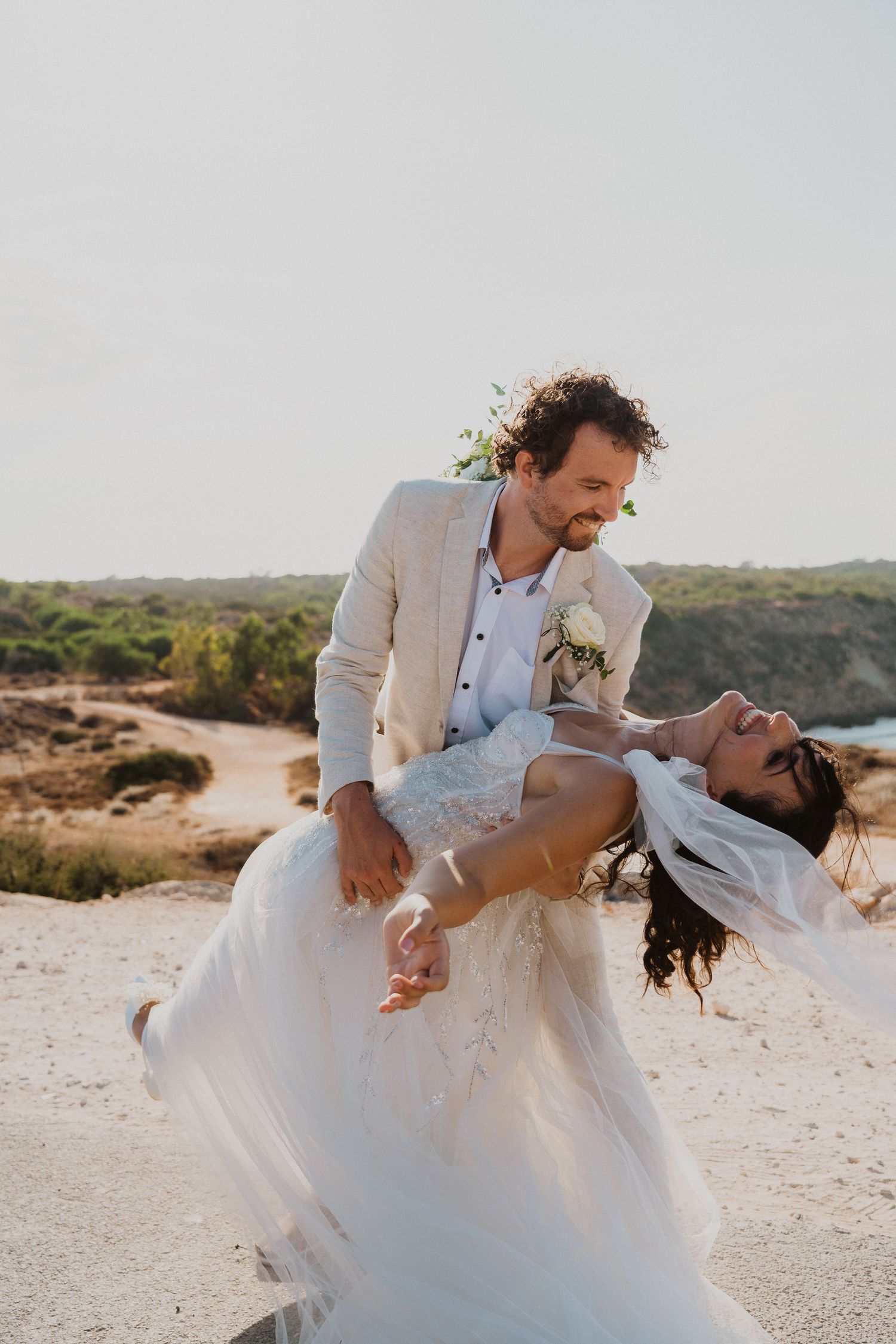 A romantic wedding couple shares a joyful dip pose against a coastal landscape at sunset.