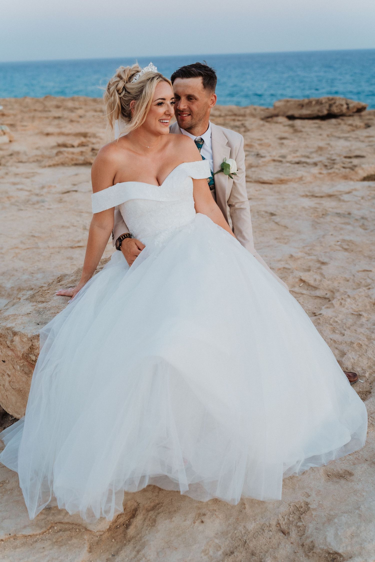 A bride in a voluminous white ball gown poses with her groom on rocky coastal terrain.