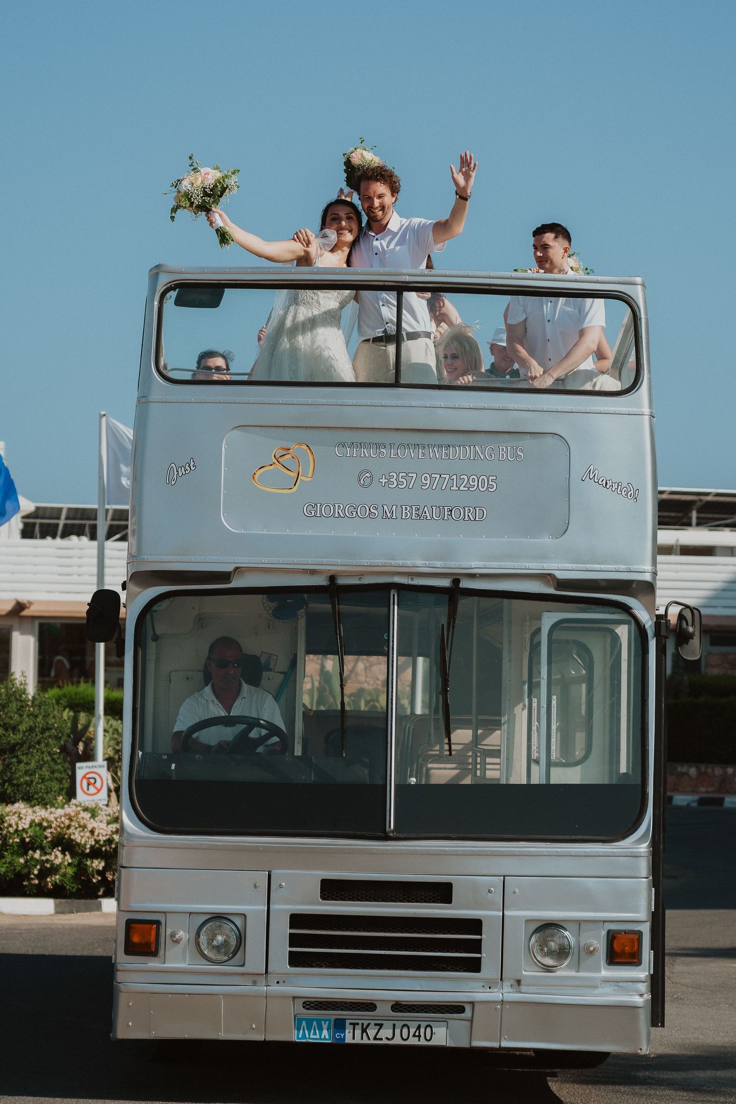 Happy couple waving from top of double-decker wedding bus with floral bouquet.