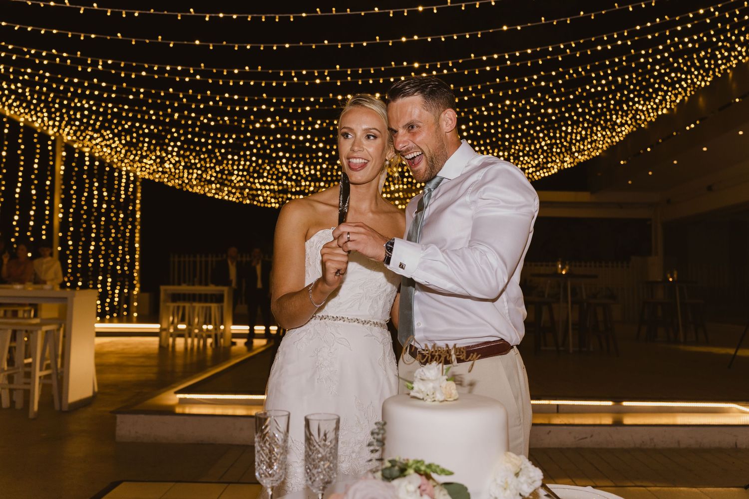 Couple shares intimate moment while cutting cake at their reception under sparkling lights.