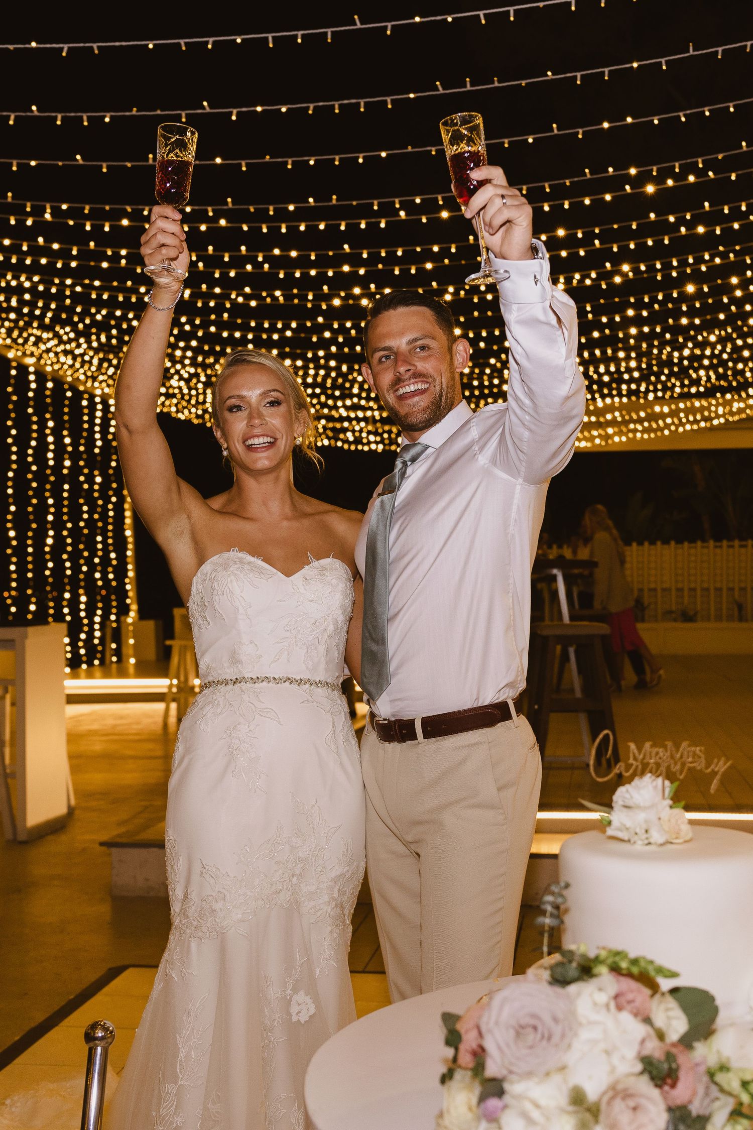 Joyful wedding couple raises their glasses in celebration under a ceiling of sparkling lights.