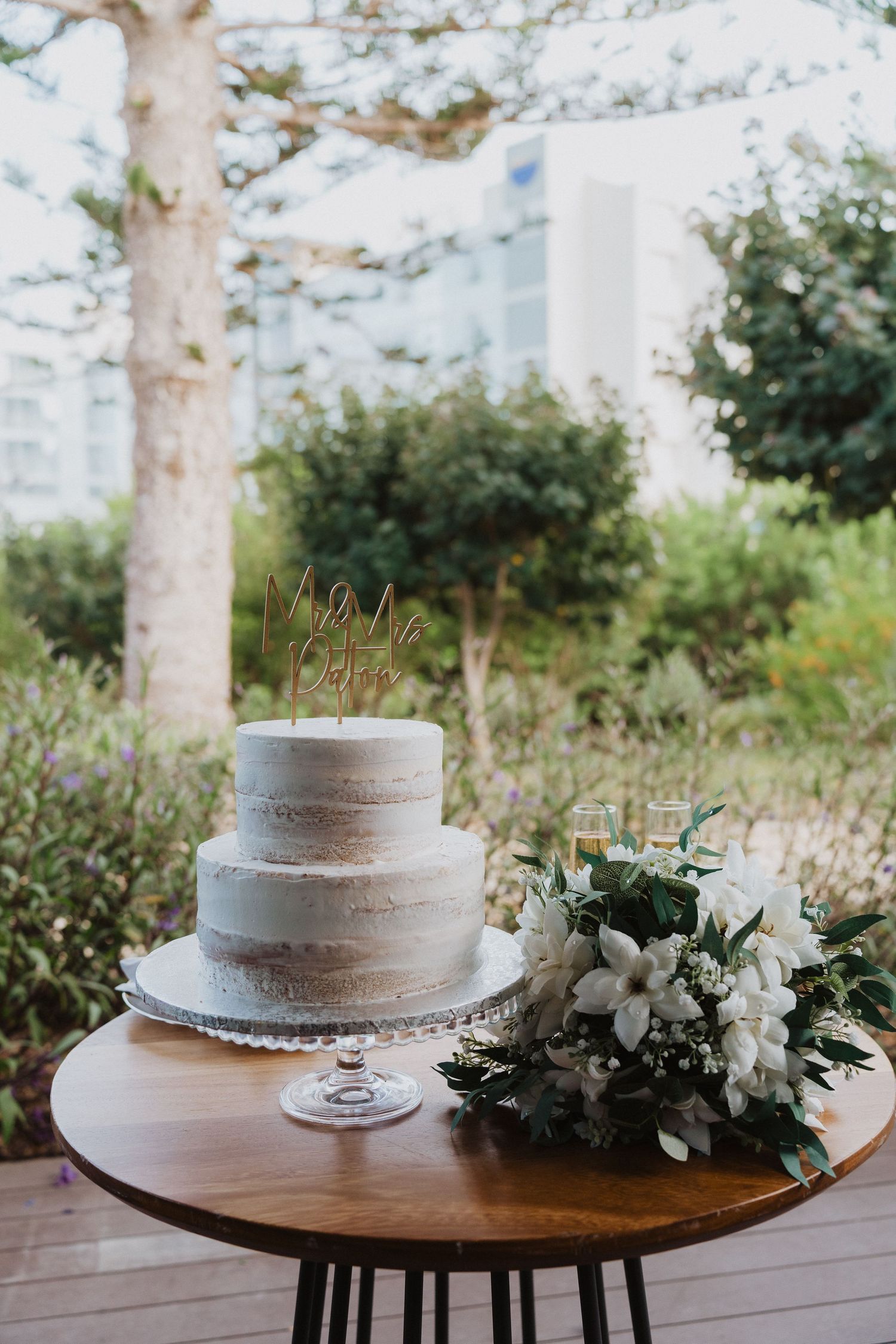 A three-tiered white wedding cake sits on a wooden table with a floral arrangement of white flowers and greenery.