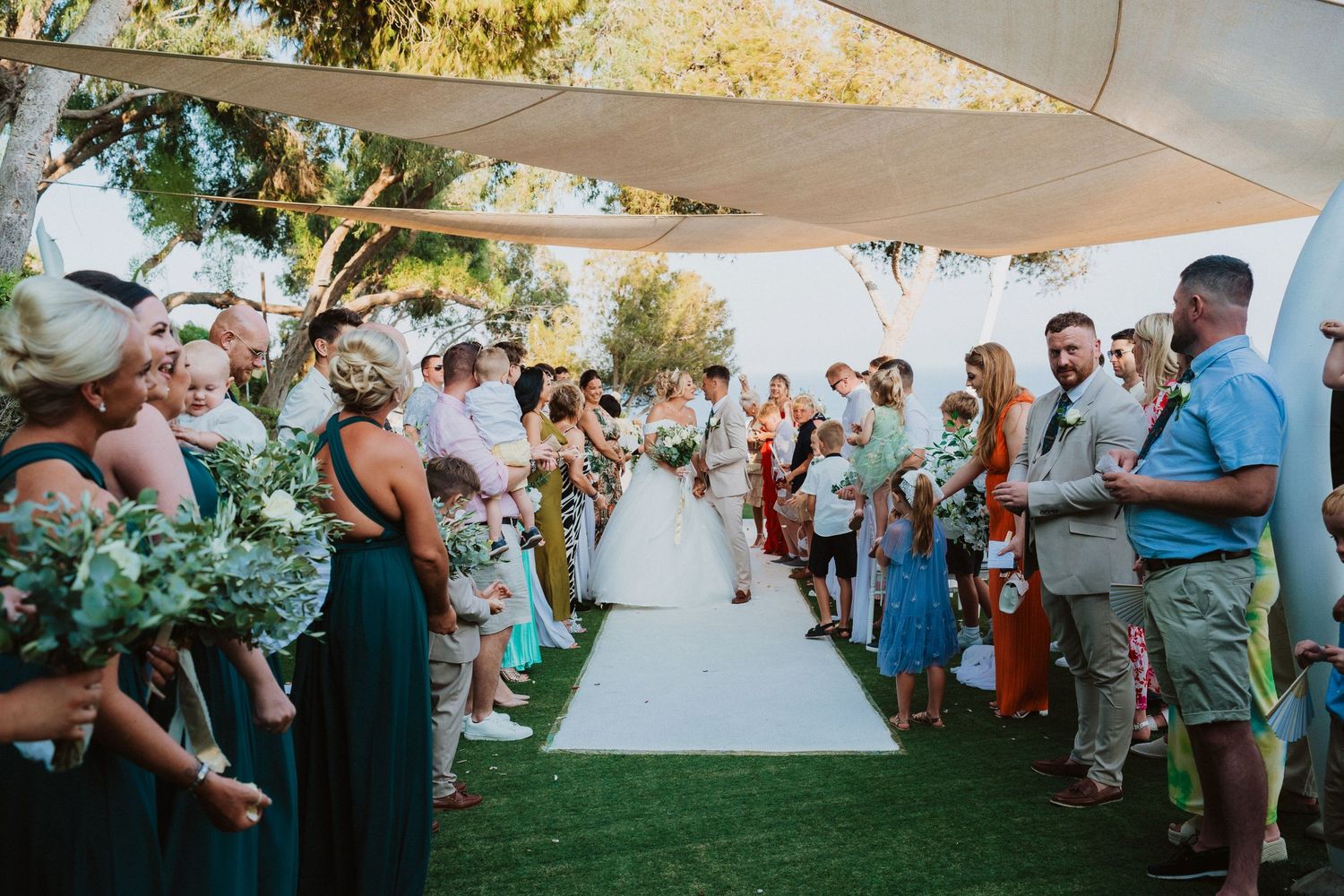 Outdoor wedding ceremony on a grassy lawn with guests lining both sides of the aisle under a white canopy.