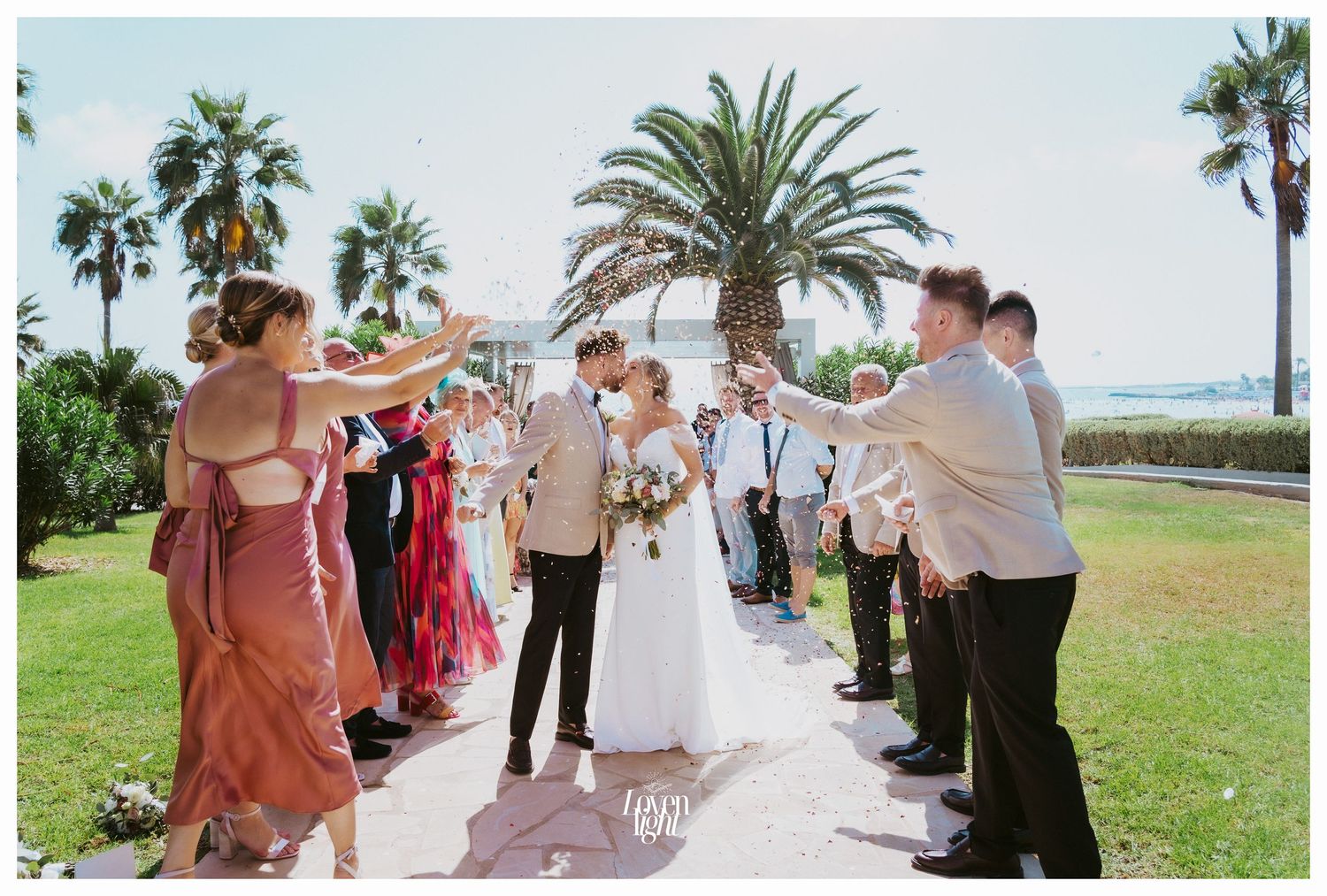 Wedding guests line a path with palm trees in the background during an outdoor celebration.
