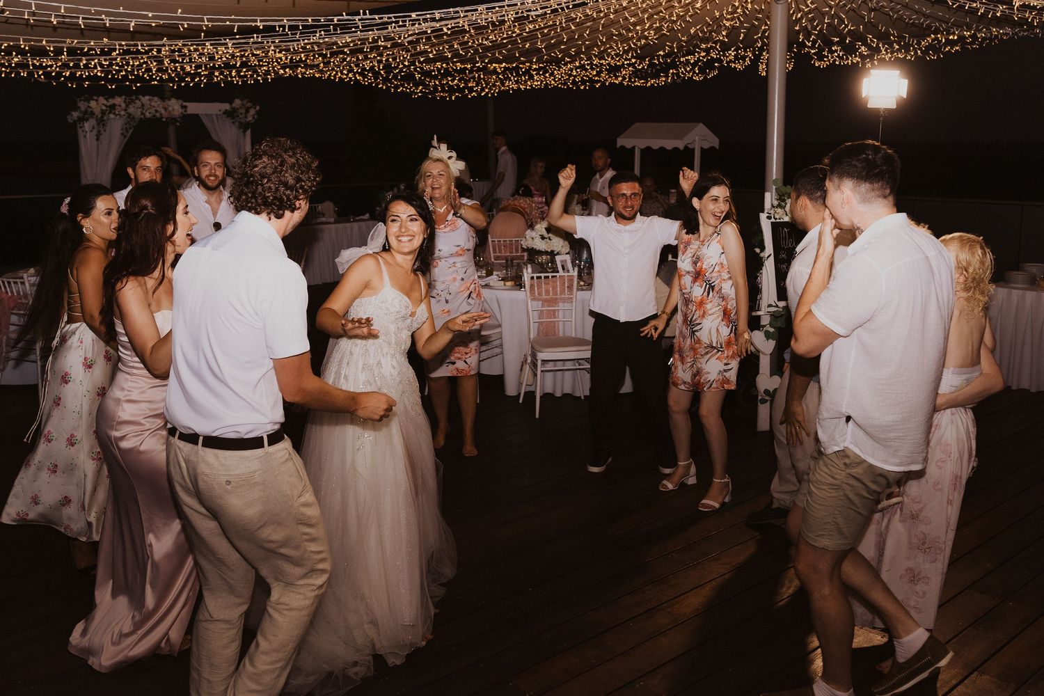 Wedding guests dance and celebrate on a festive dance floor illuminated by overhead string lights.